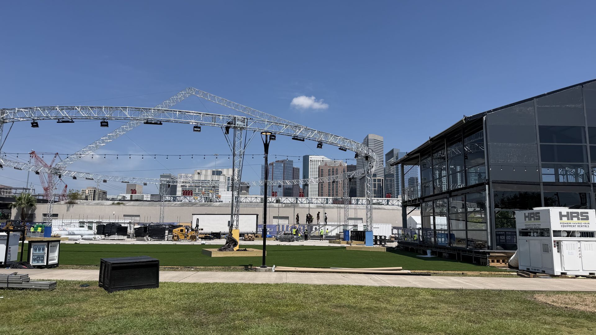 Outdoor construction site under a clear blue sky, with large metal truss scaffolding forming arches, a glass-front building to the right, and a city skyline in the distance over a grassy lot.