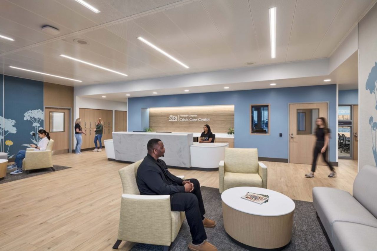 Lobby of a Crisis Care Center with blue walls, light wood floor, and a white curved reception desk. A man sits in a beige chair while others walk; a sign behind the desk reads "Crisis Care Center".