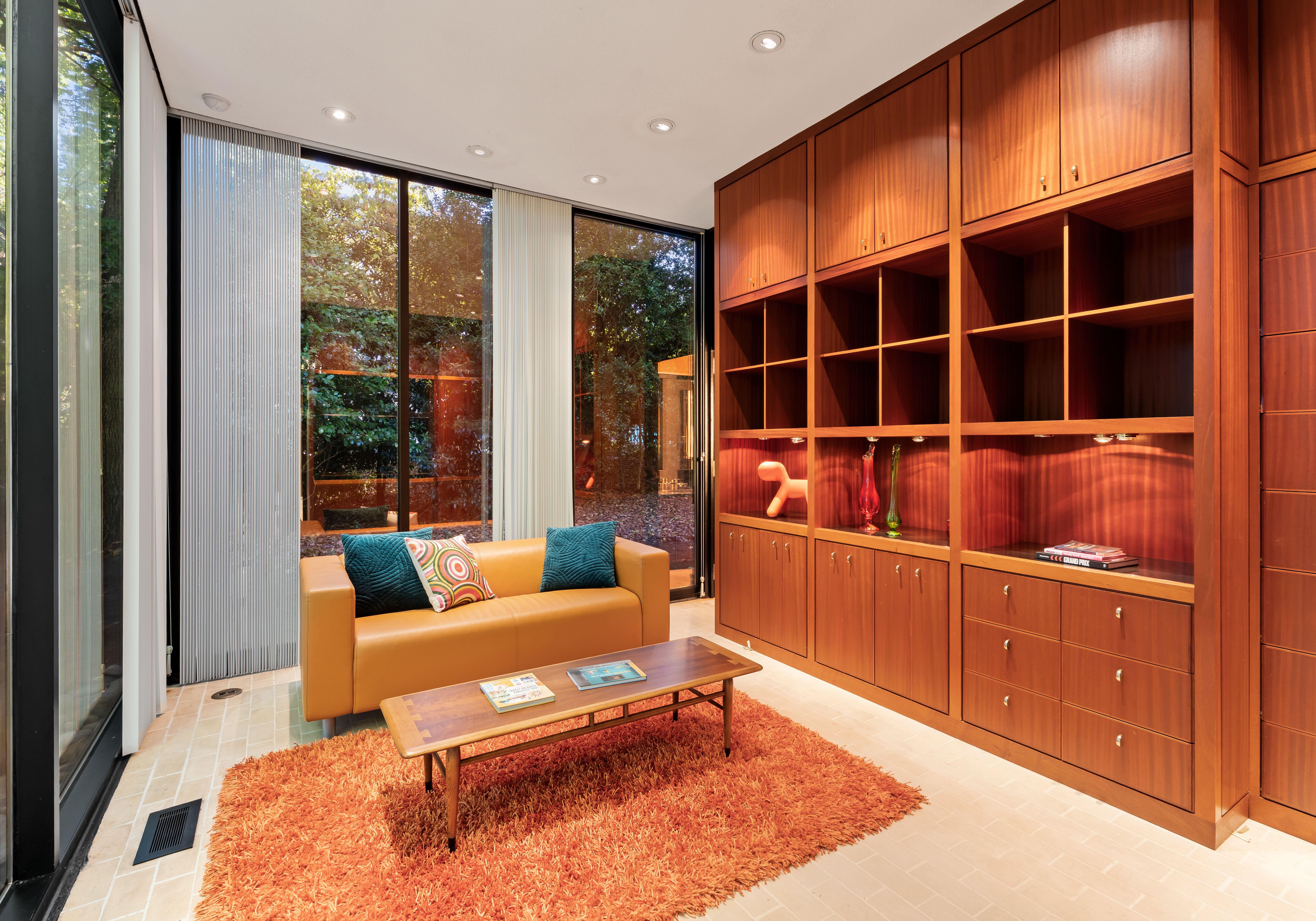 Modern living room with large wooden shelves, a mustard-yellow sofa with teal and patterned pillows, a wooden coffee table, and an orange shag rug, next to floor-to-ceiling windows.