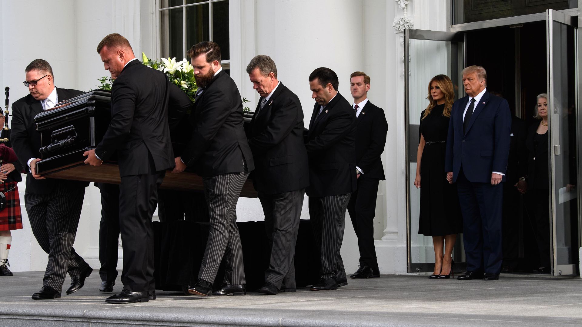 Donald Trump and Melania Trump stand in a doorway as a casket with white lilies on top is carried away 