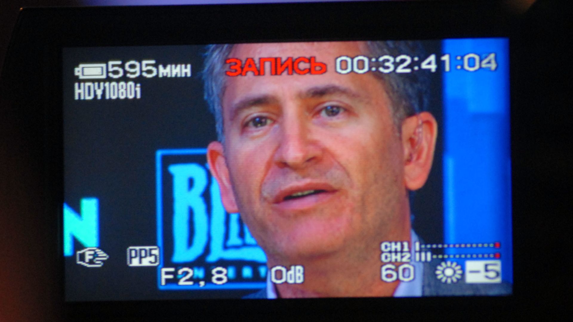 Photograph of a man speaking, as he is seen through the viewfinder of a video camera