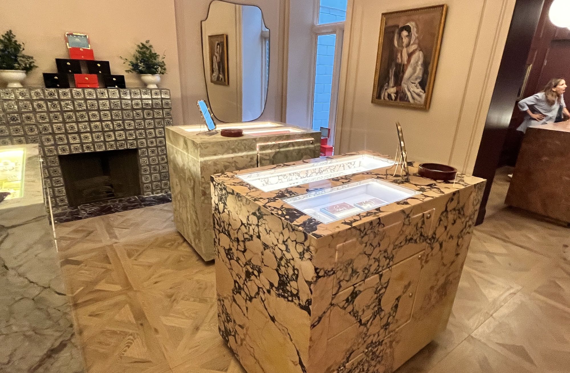 Luxurious room with a marble island in the foreground, a tiled fireplace on the left, a tall mirror and a painting on the wall, potted plants, and a woman leaning at a wooden desk on the right.