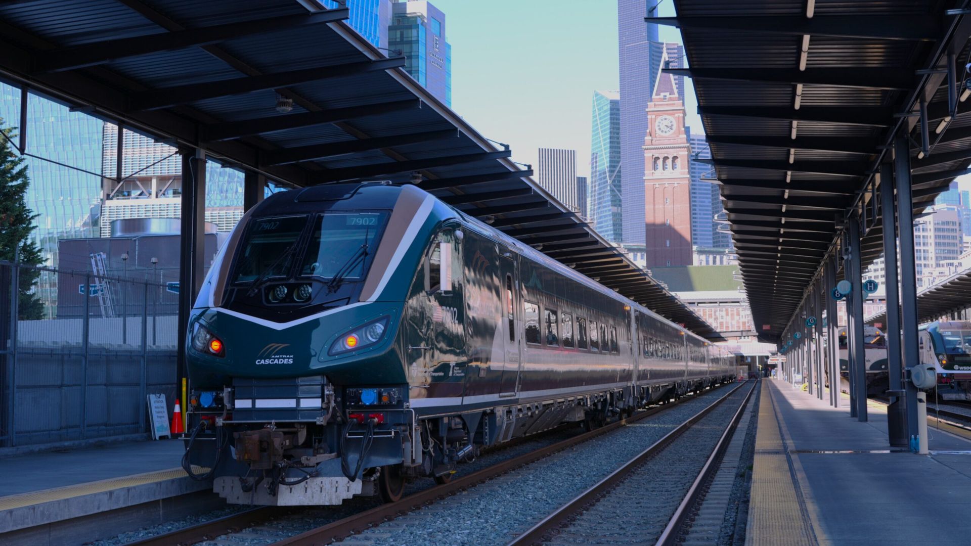 Teal-blue Amtrak Cascades locomotive at a city train platform with dark metal canopies; glass skyscrapers and a brick clock tower visible in the background under a bright blue sky.