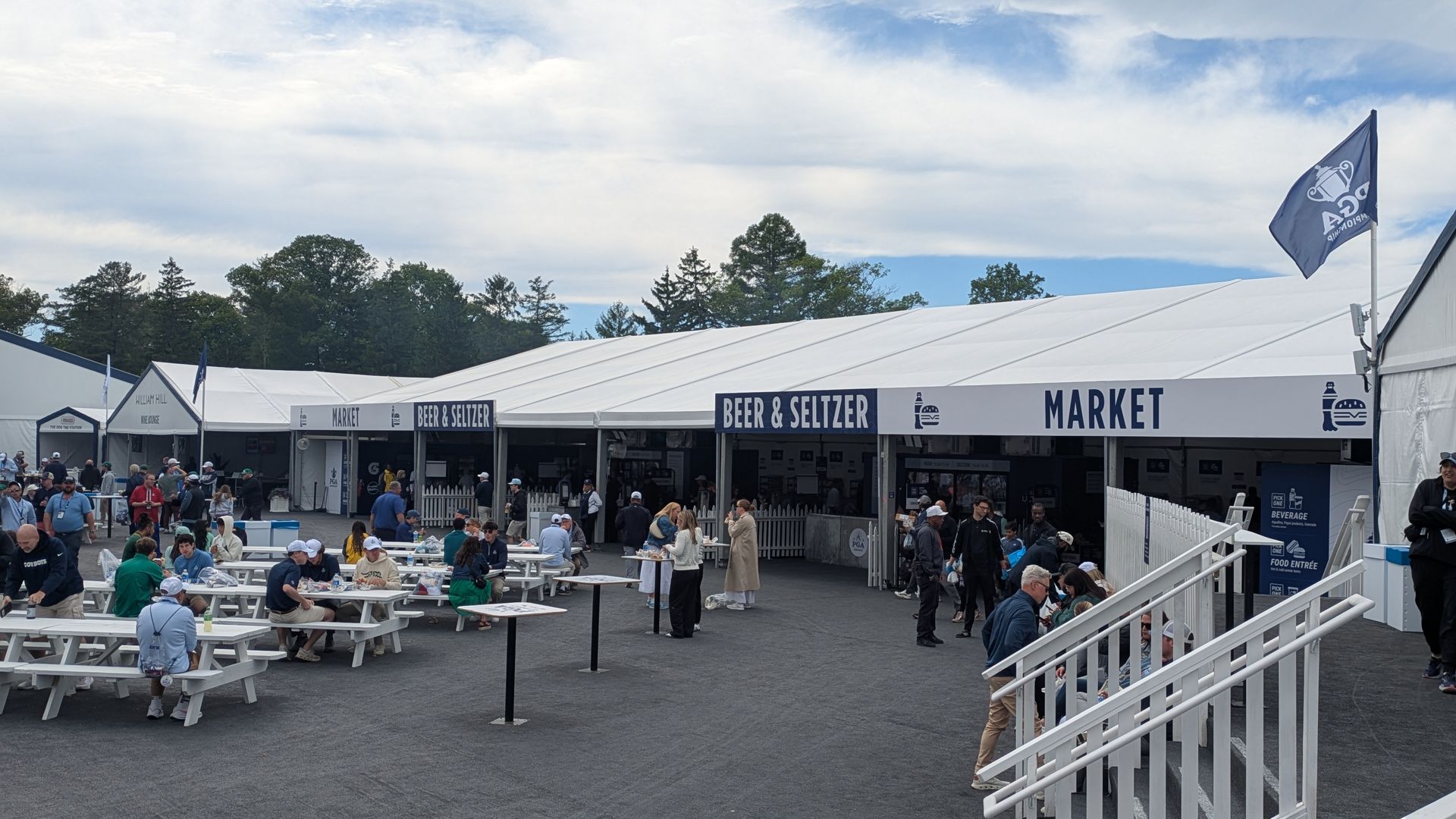 Outdoor festival with white tents labeled "BEER & SELTZER" and "MARKET." People sit at white picnic tables and walk among stalls; a blue flag waves as trees and a blue sky backdrop looms.