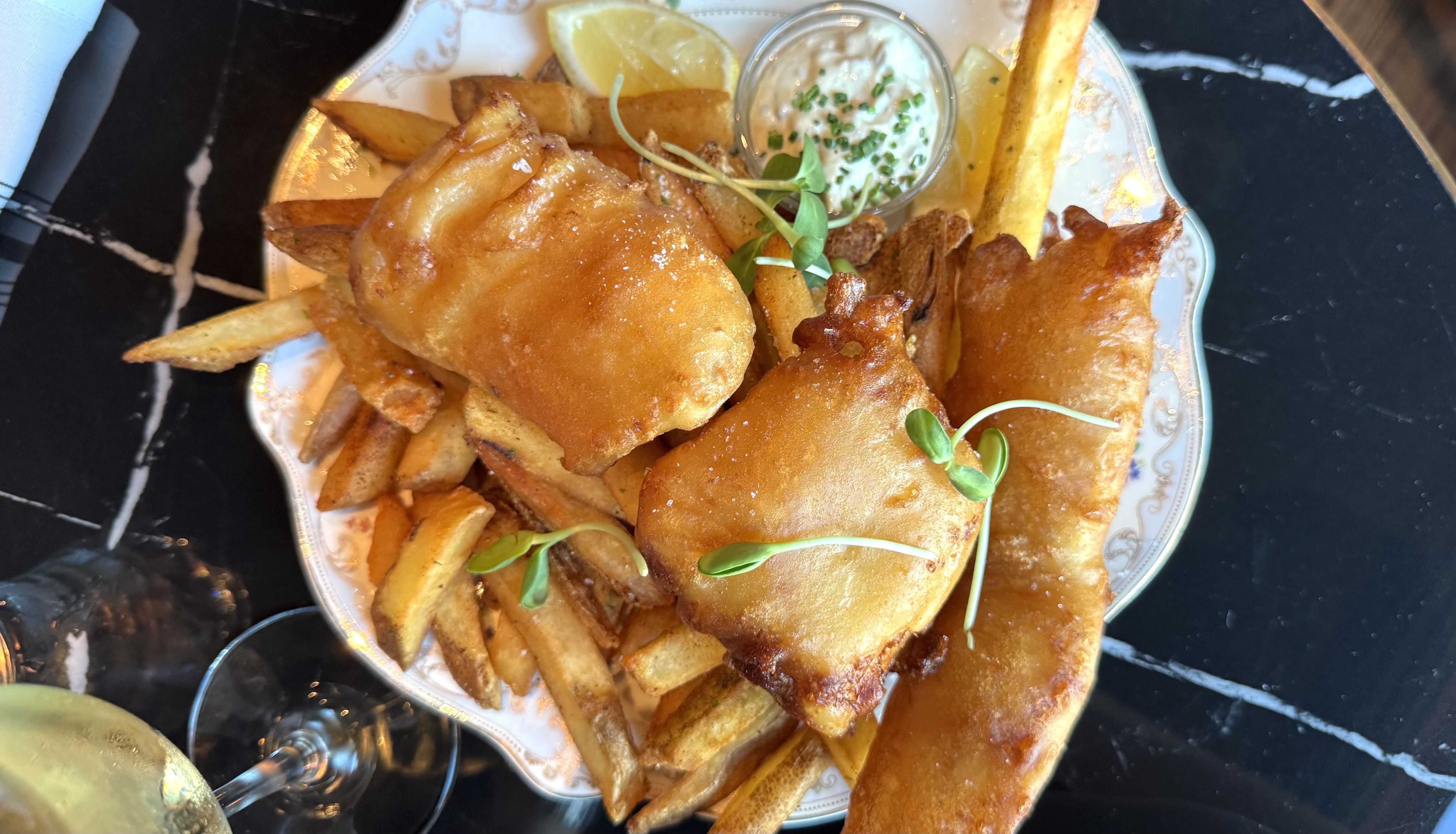 Overhead view of a plate of golden fried cod pieces and thick potato fries, with a small cup of creamy tartar sauce and chives, lemon wedges, and microgreens on a decorative plate.