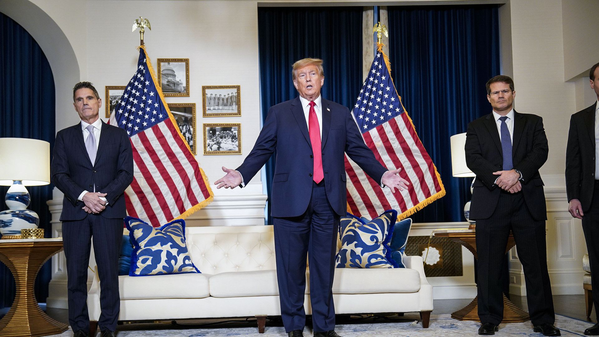 Former President Trump stands with his hands outstretched, standing in front of two large American flags and a white couch, while speaking to the press in a D.C. hotel room.