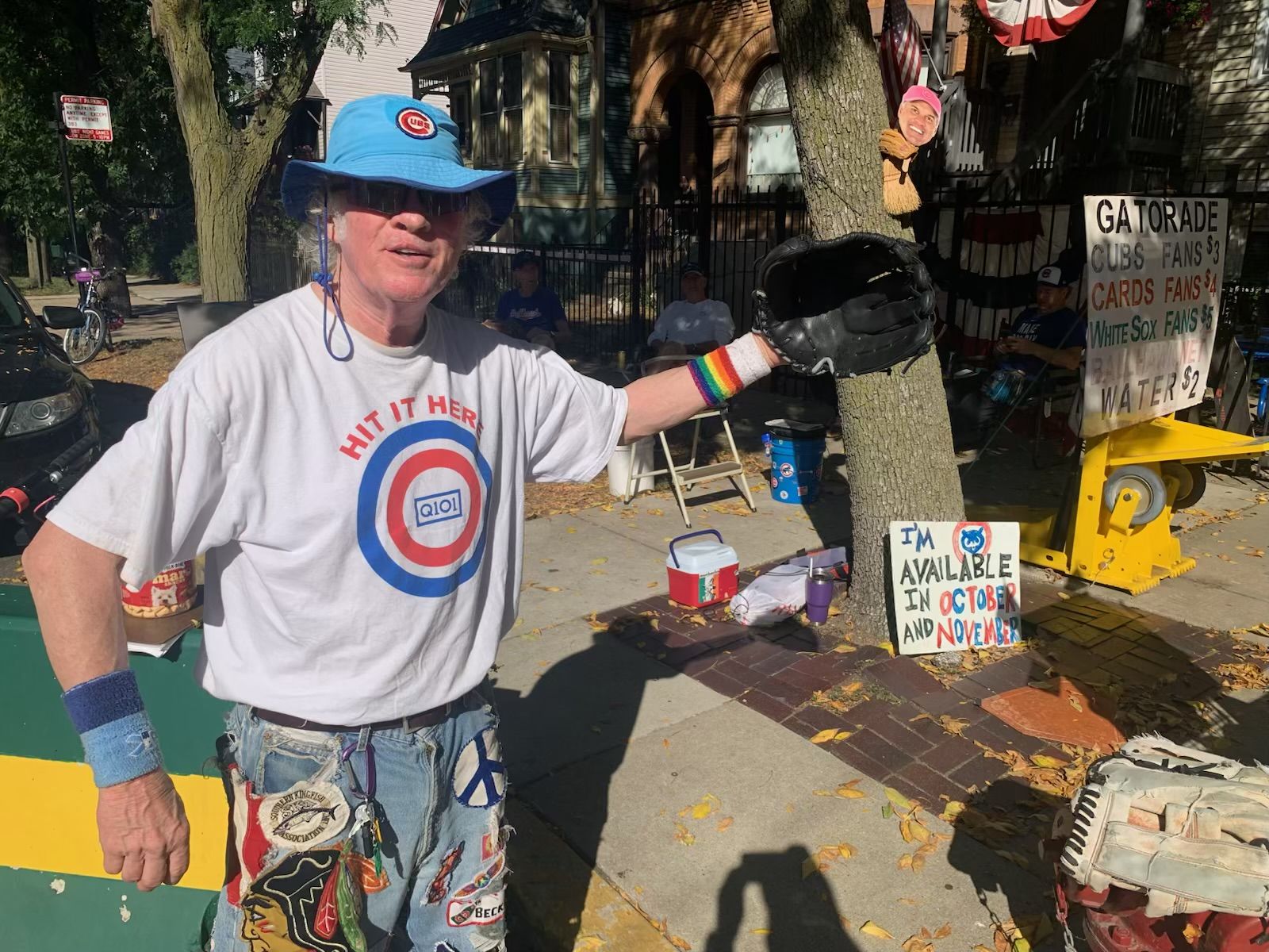 Scott Doherty wearing a blue Cubs bucket hat, white "Hit It Here" T-shirt, rainbow wristband, and denim shorts with patches, holding a black baseball glove on a sidewalk with signs behind him.