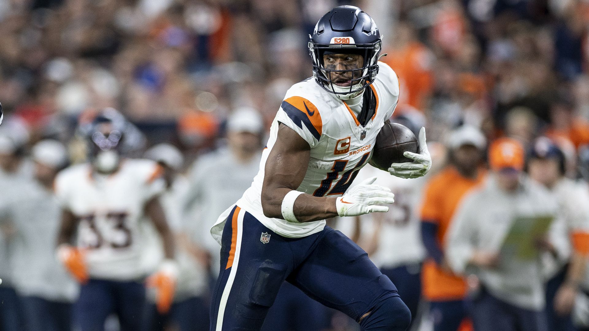 A man in a navy helmet and white jersey holds a football closely while running.