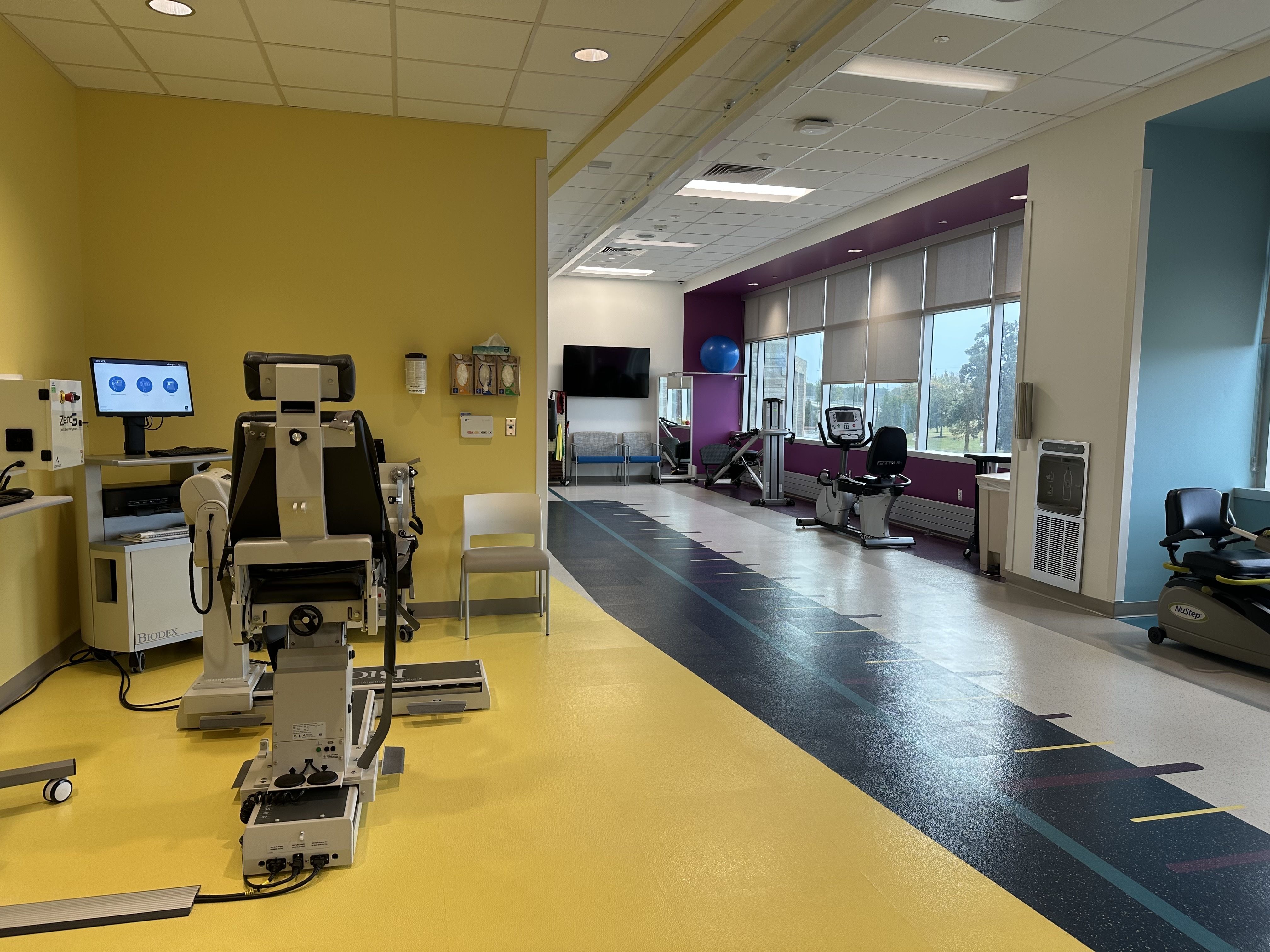Bright rehab gym with a yellow left wall, foreground exercise machines, a purple accent wall by large windows with blinds, and a blue ball on a purple pillar; cardio machines along the windows.