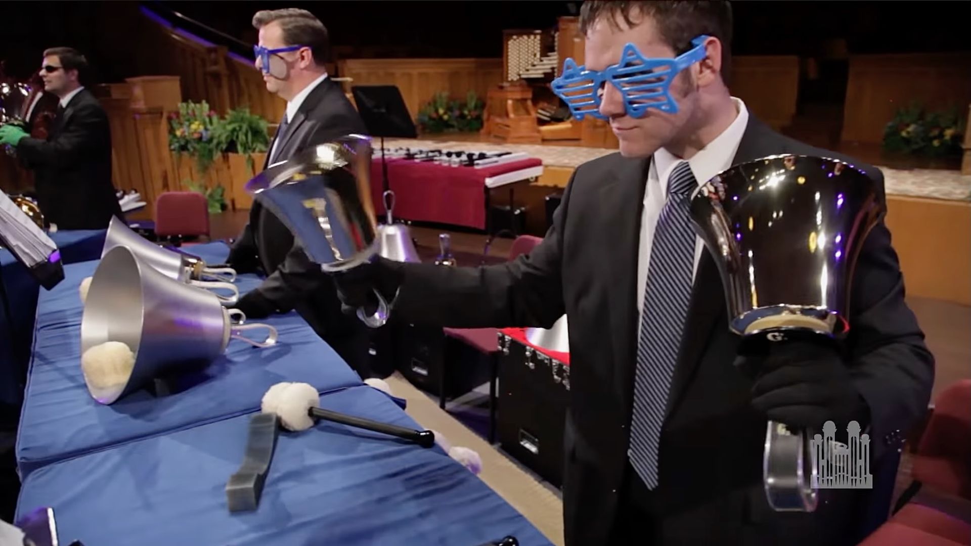 Men in sunglasses play handbells over a blue padded table.