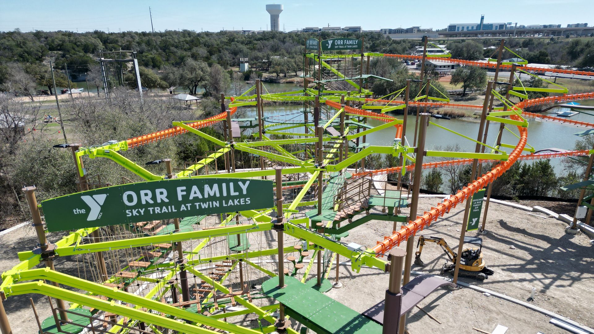 Bird's-eye view of a bright lime-green and orange YMCA sky trail course under construction, with wooden platforms and rails near a lake and trees, titled "Orr Family Sky Trail at Twin Lakes."