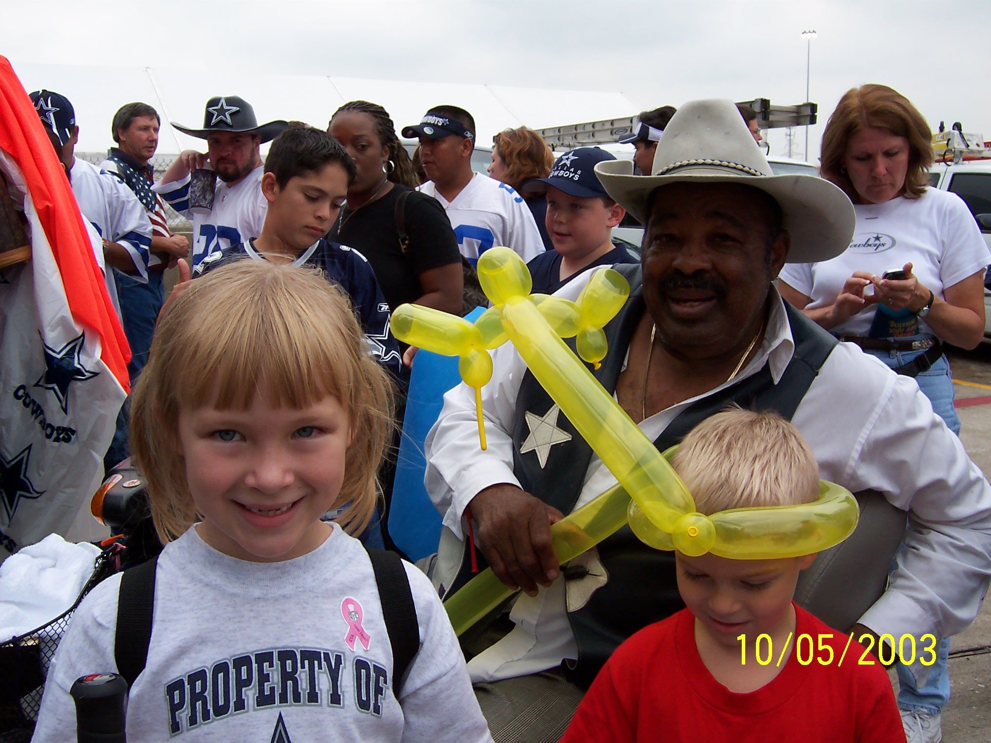 Group of people, many wearing Dallas Cowboys shirts and hats. Older man in cowboy hat holds yellow balloon animals, one child wears balloon hat. Date stamp 10/05/2003.