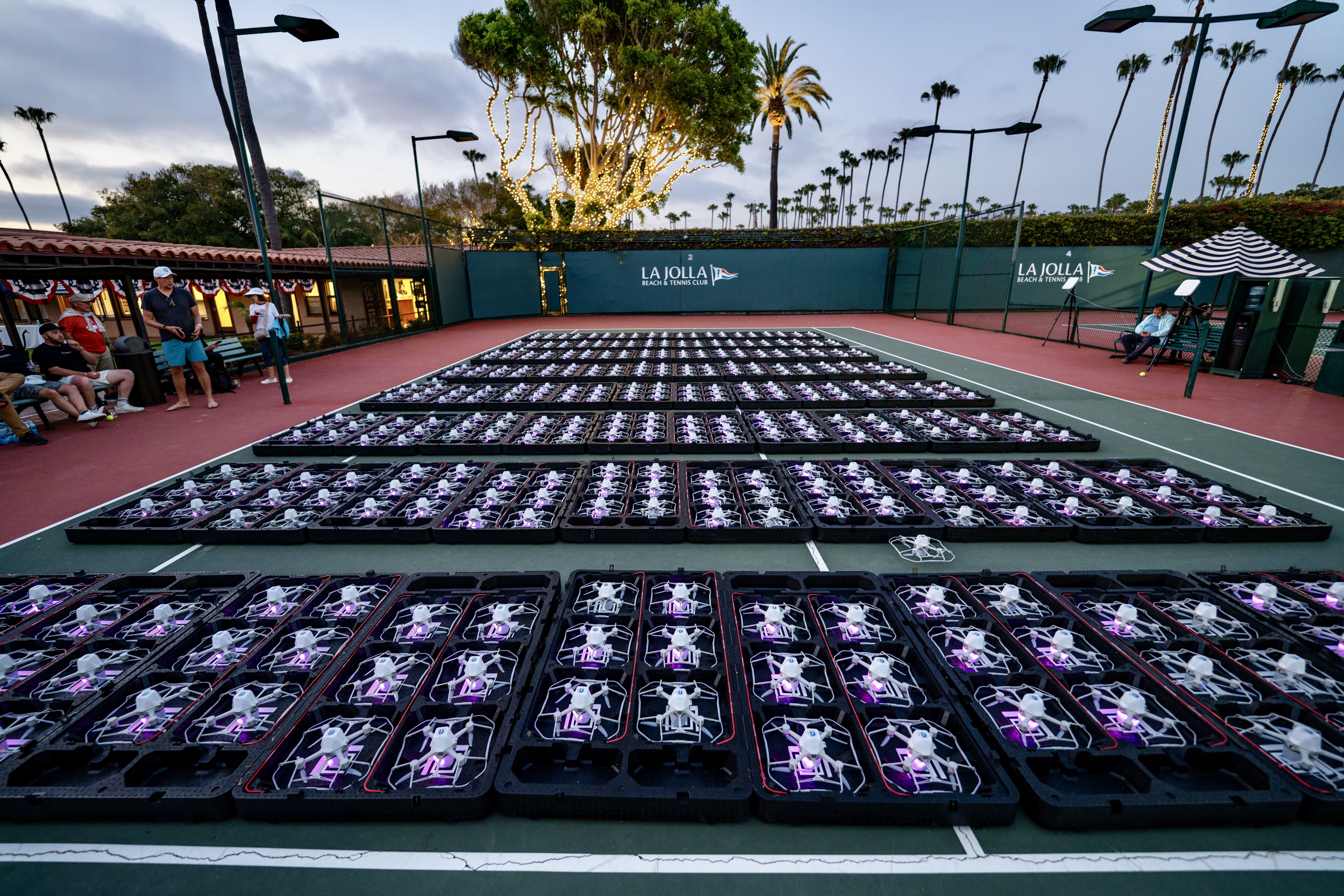 Wide view of a tennis court at La Jolla Beach & Tennis Club, filled with rows of black trays holding purple-lit items. Left: people by a building; center: tree with lights; right: striped canopy and palms.