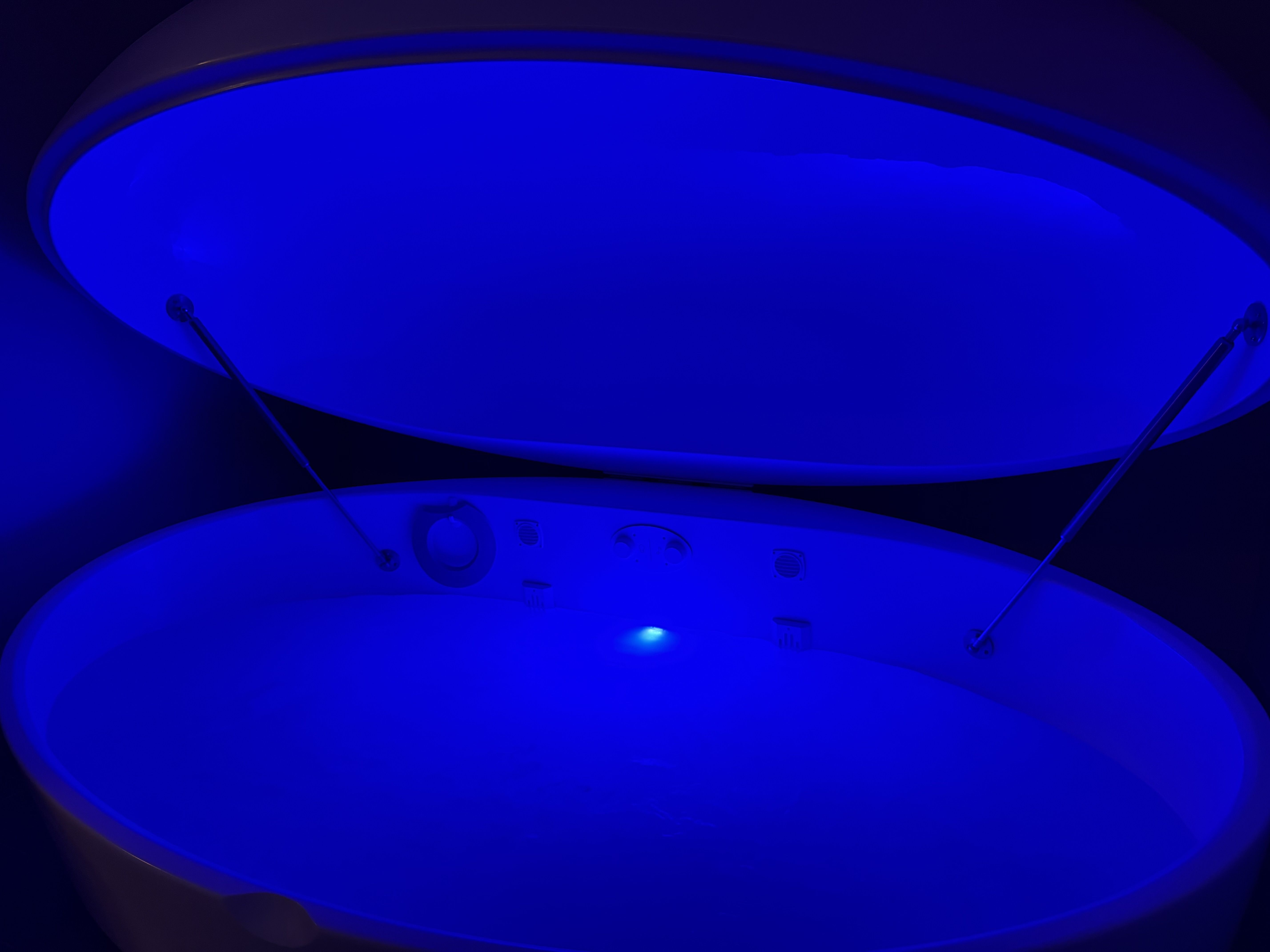A large white sensory deprivation float tank illuminated by blue light, with its lid open and water inside.