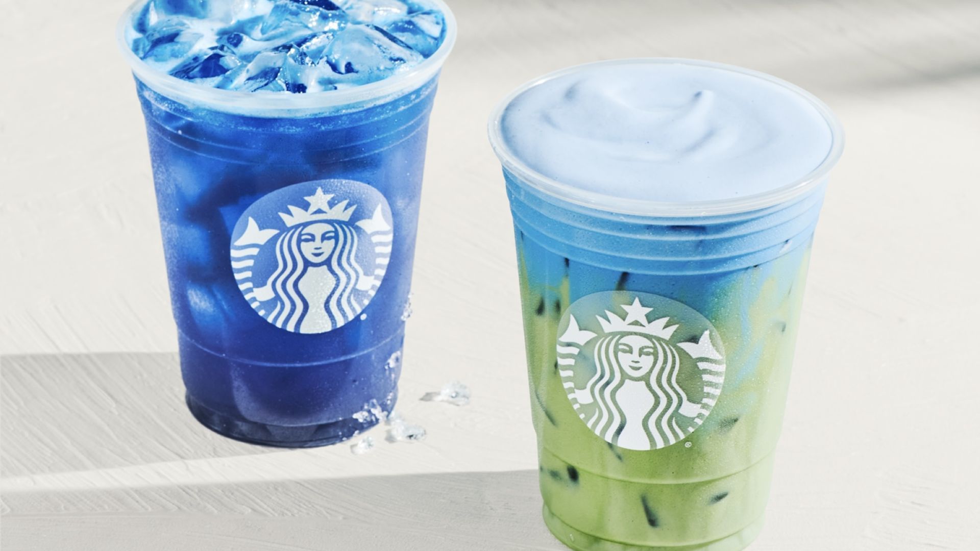 Two Starbucks cold drinks in clear cups on a light wooden table; left cup dark blue with ice, right cup blue-to-green gradient with ice; both display the white Starbucks logo.