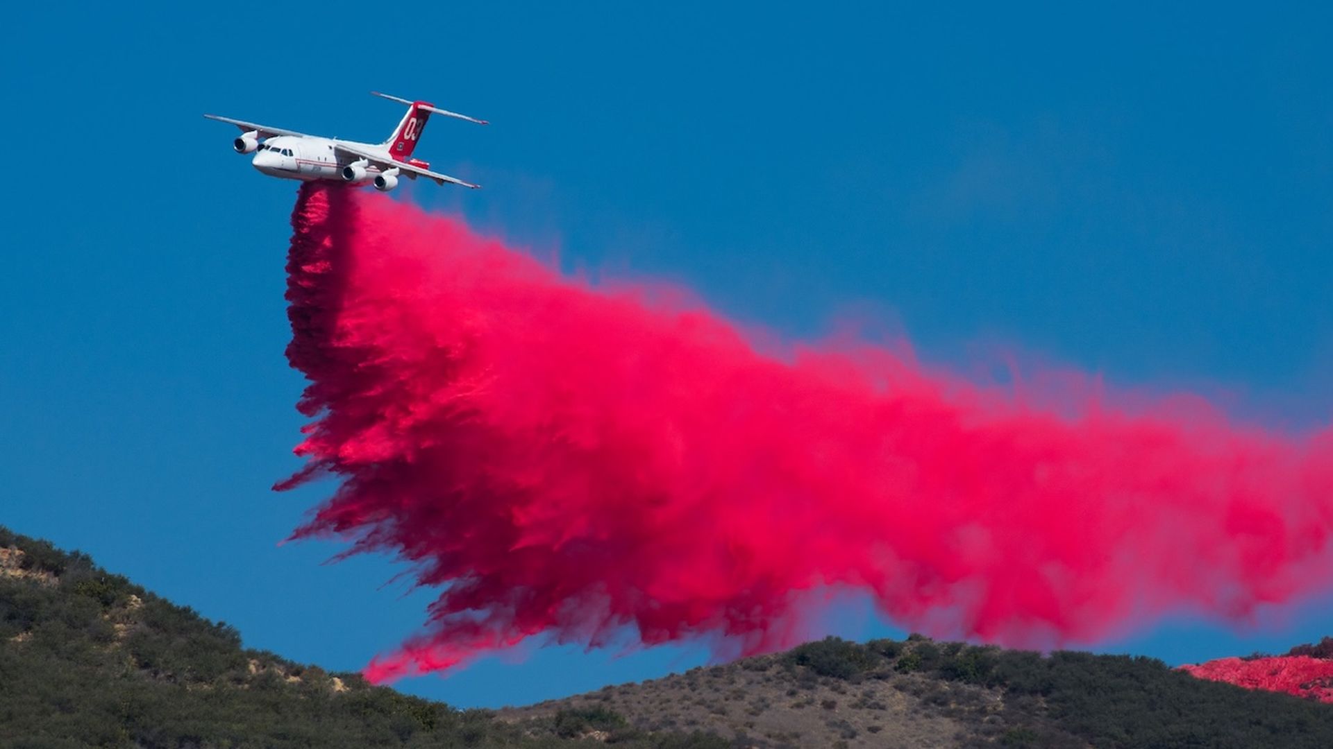White aircraft with red tail releases bright red smoke trail as it flies above hills, creating a thick crimson plume against a clear blue sky.