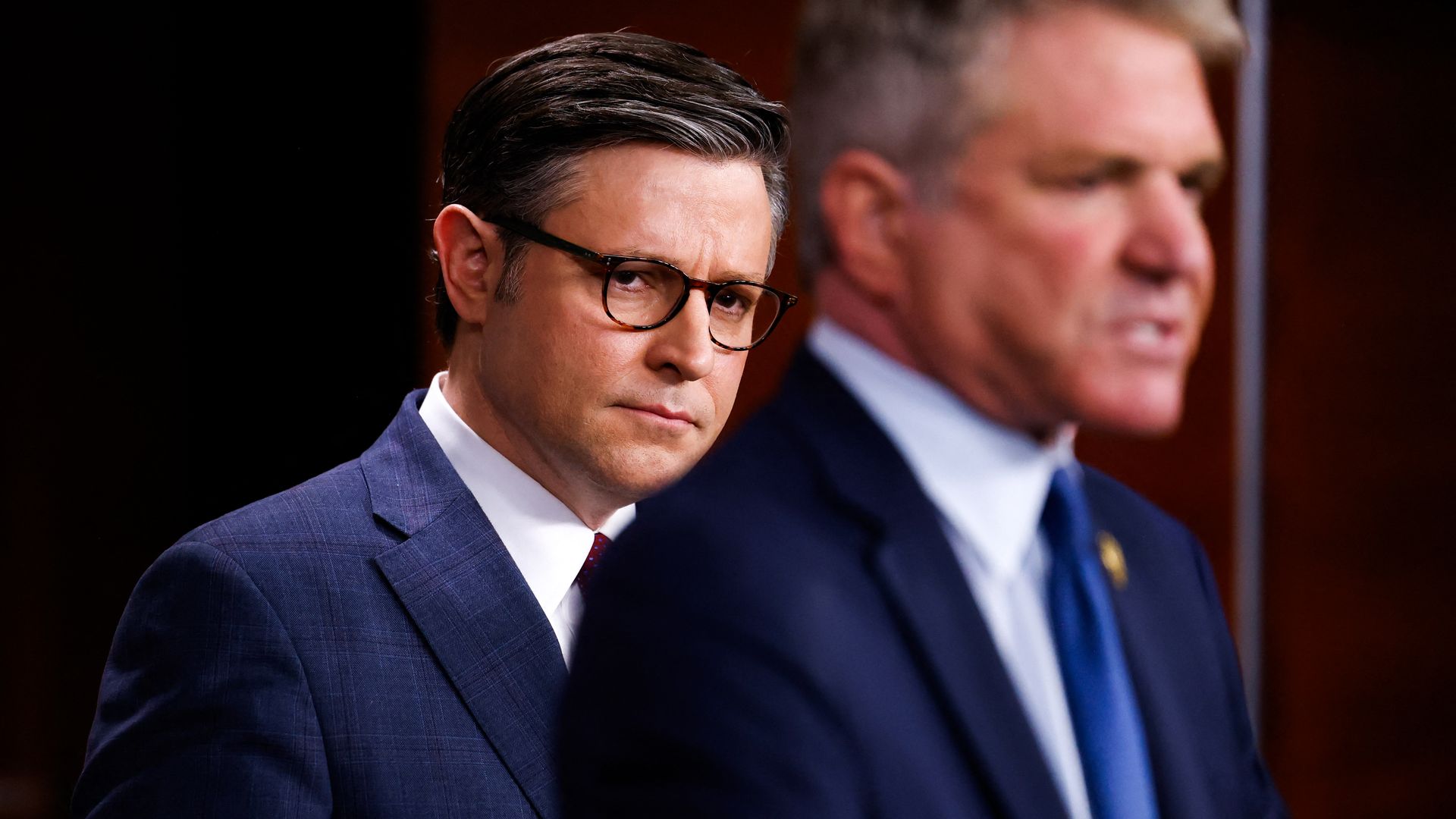 House Speaker Mike Johnson, wearing a blue suit and standing behind Rep. Michael McCaul in the wood-paneled House TV studio.
