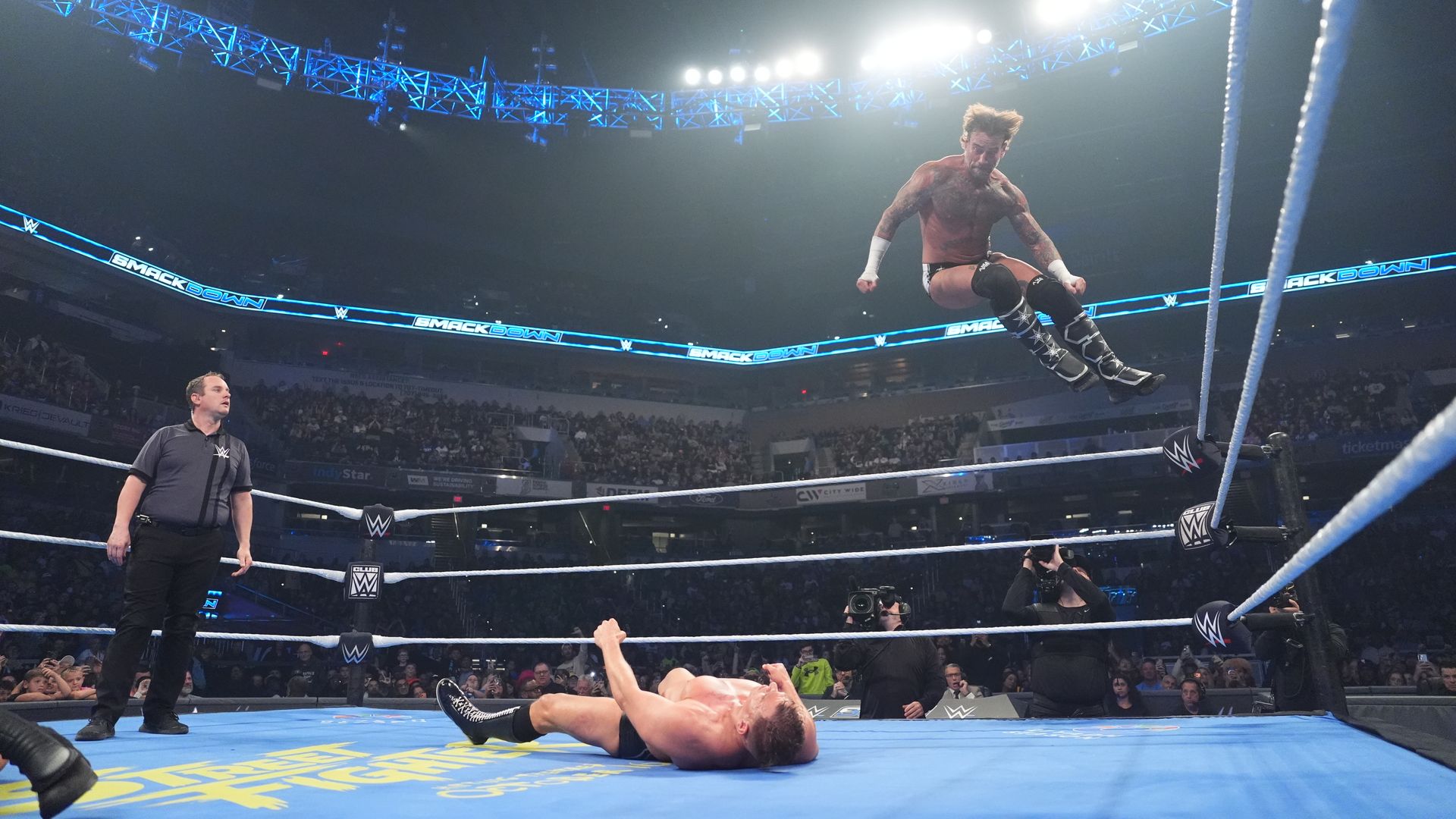 A wrestler is photographed in midair as he appears to jump toward another wrestler lying prone on the floor of a wrestling ring.
