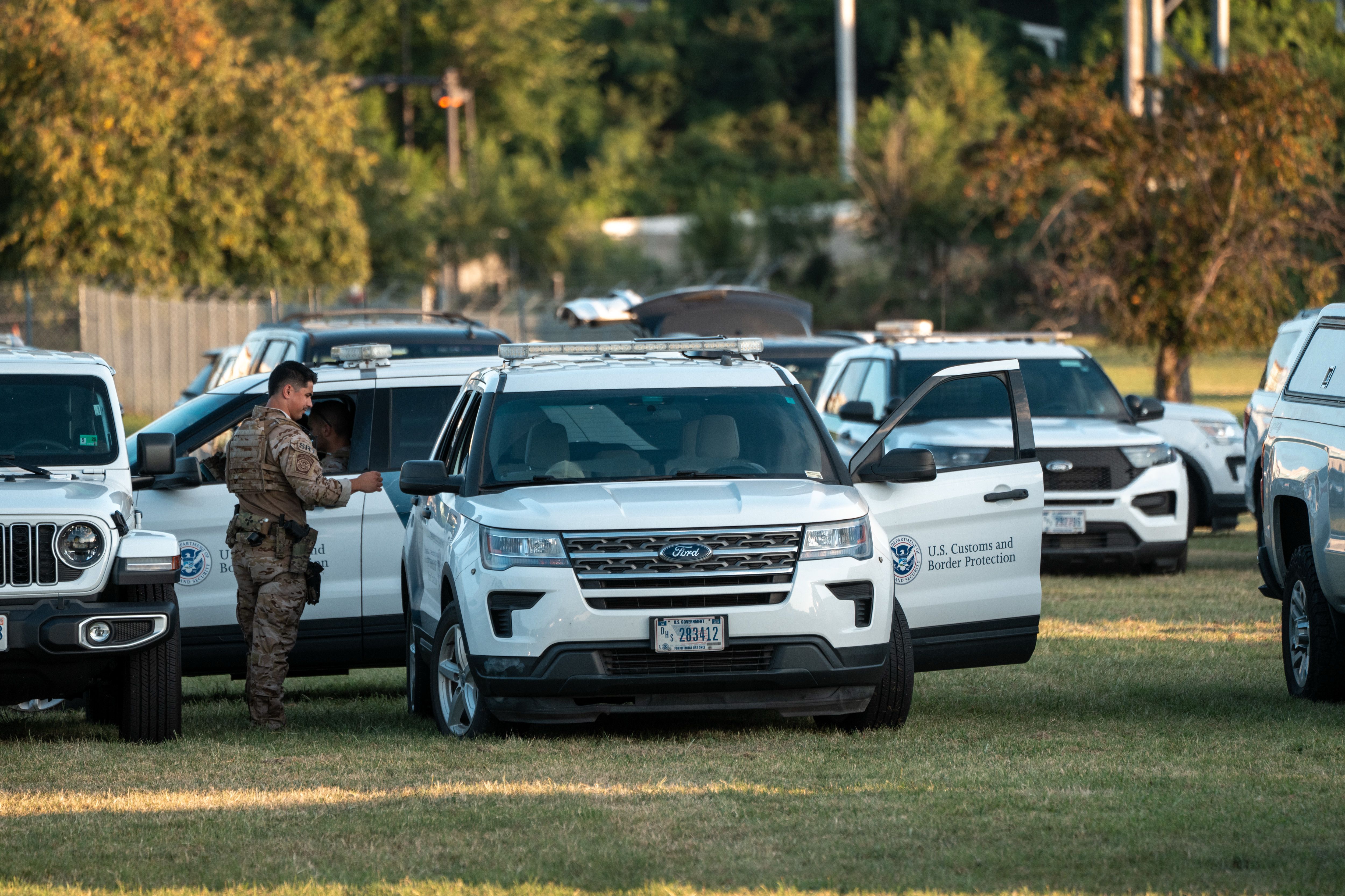 U.S. Customs and Border Protection officers in tan uniforms stand near white marked SUVs in a grassy area with trees in the background during daylight.