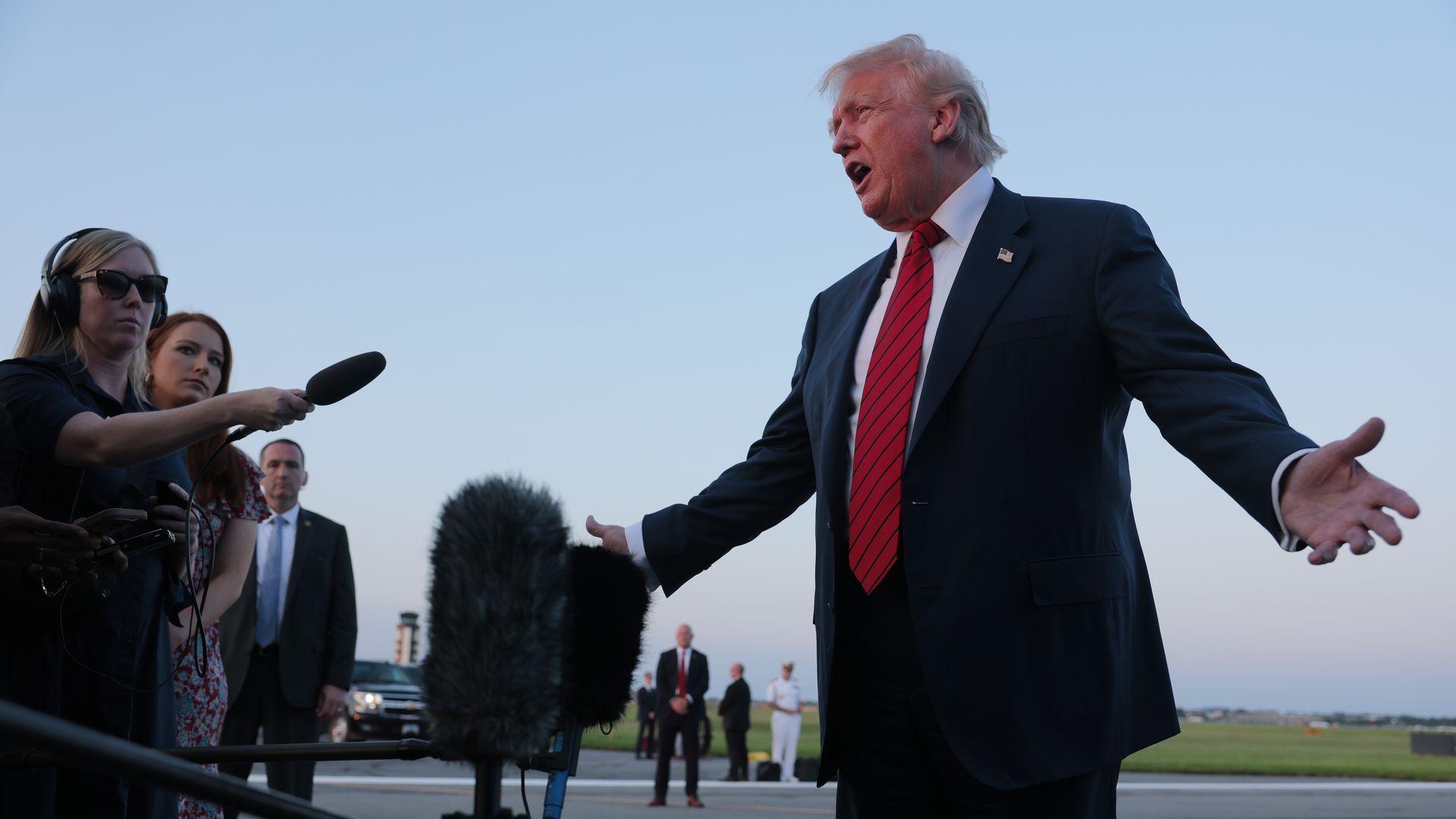President Trump, in a dark suit, white shirt, and red striped tie speaks with arms open outdoors on an airfield during sunset, while reporters record him with microphones.