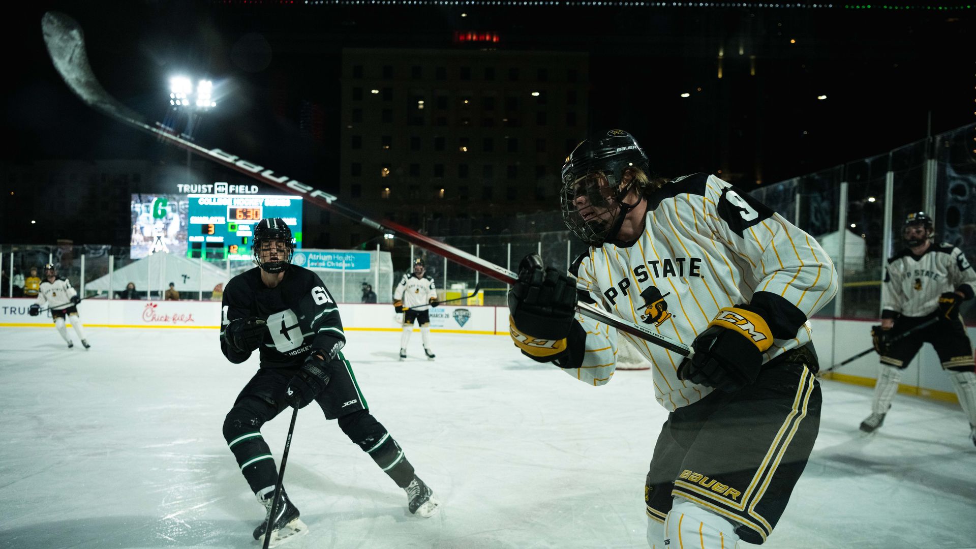 Ice hockey game at night with players in action; one in white and yellow pinstriped PP State jersey and another in black jersey with number 61, illuminated rink with scoreboard in background.