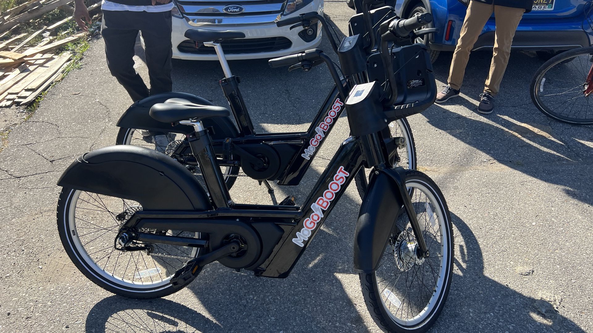 Two black e-bikes with the red MoGo logo are shown next to each other in front of people and a couple cars.