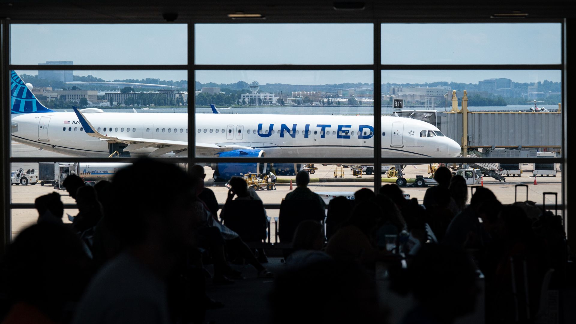 Silhouette of people sitting in an airport terminal with a United Airlines plane parked at gate B11 outside the large windows under a clear sky.