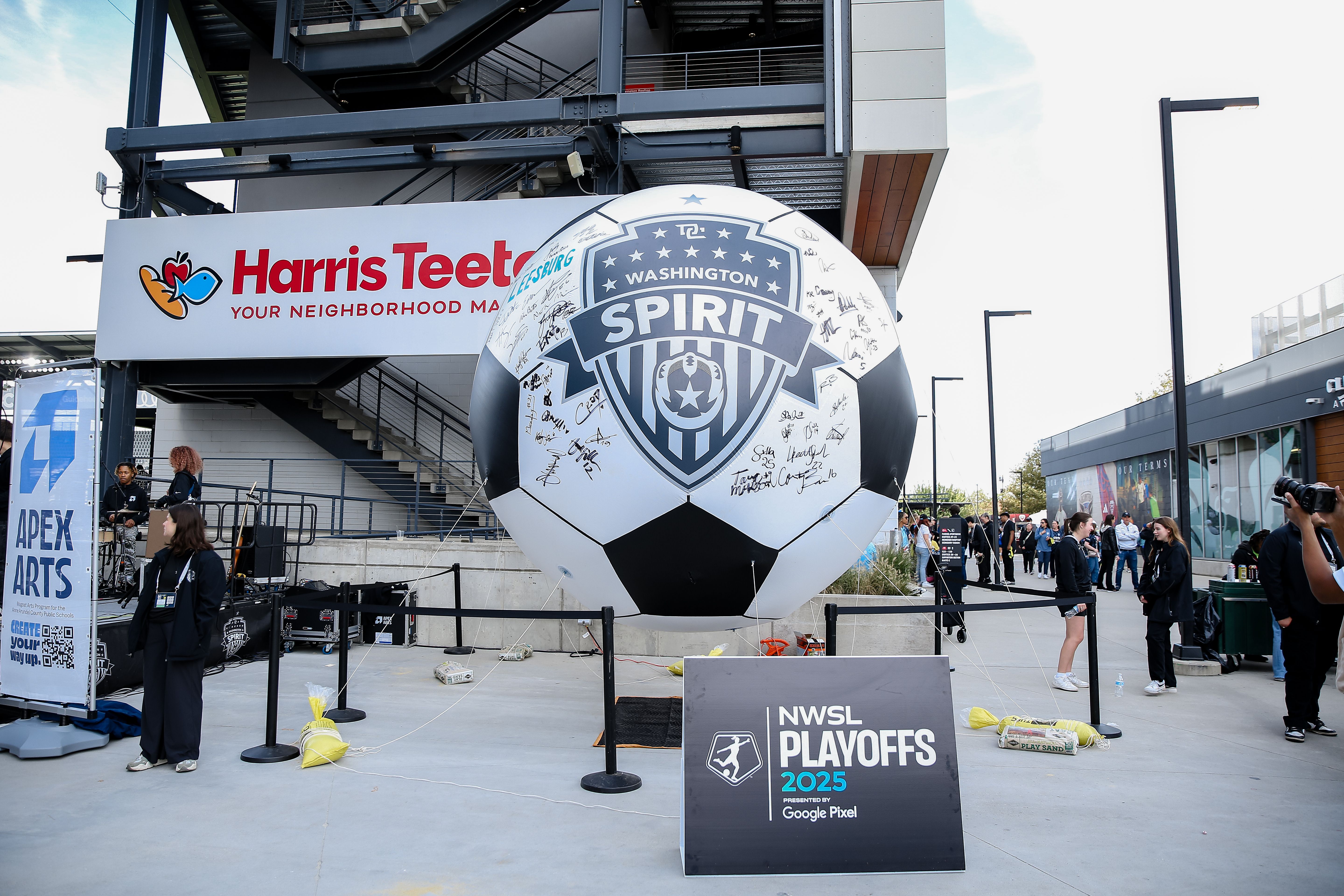 Large black and white soccer ball with Washington Spirit logo and signatures in front of Harris Teeter sign at outdoor event with NWSL Playoffs 2025 sign and people around.