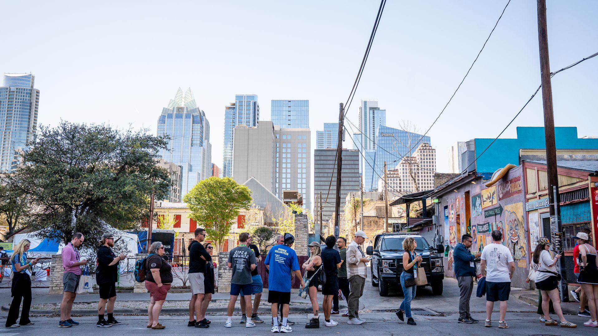 A line of people stands along a city street, graffiti walls on the right, a black SUV parked nearby, and distant glass skyscrapers under a clear blue sky.