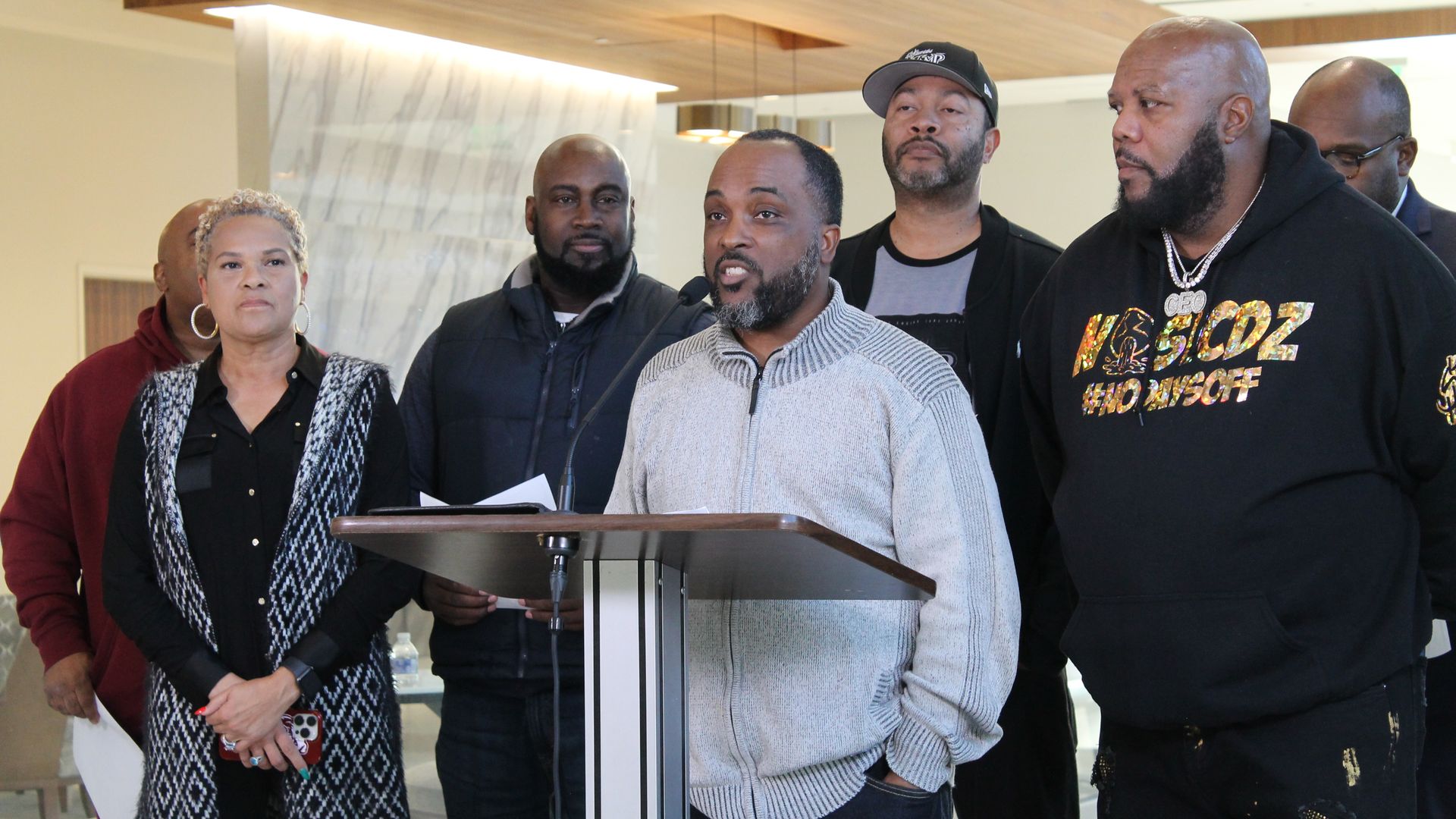 Group of six people standing indoors; man in gray sweater speaking at a podium with microphone, others stand behind him listening attentively in casual clothing.