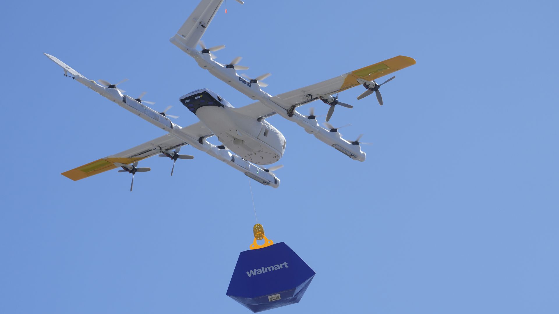 White multi-rotor delivery drone flies against a blue sky, lowering a blue Walmart cargo box attached by orange/yellow rigging.