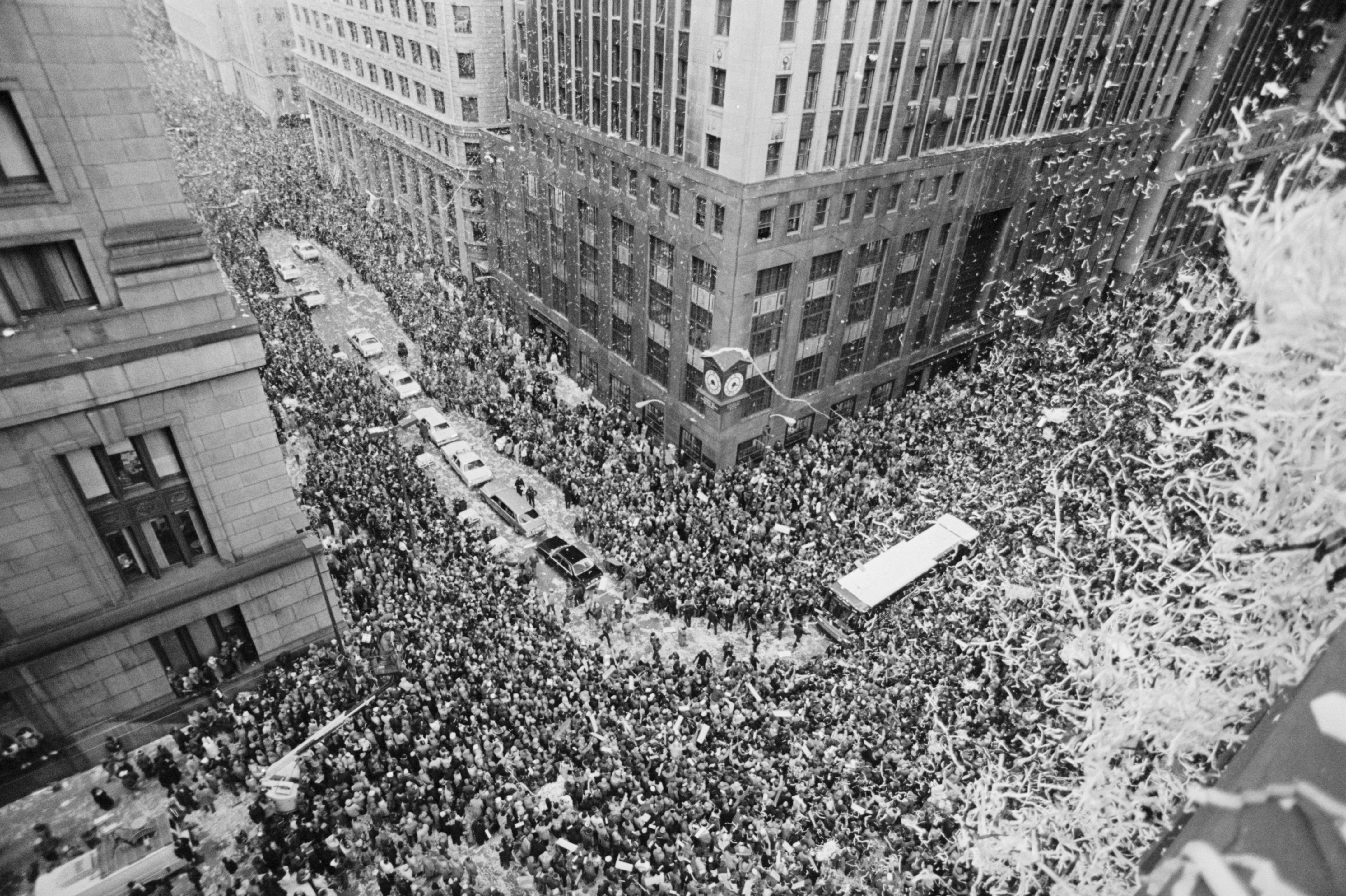 Black and white aerial photo of a massive crowd filling a city street between tall buildings, with cars and a bus visible, and confetti raining down on the scene.