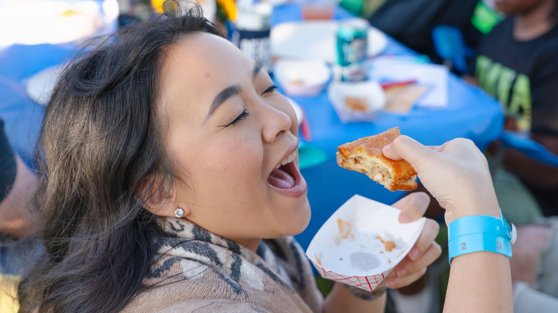 Woman with long dark hair and a beige sweater eagerly eating a bitten beignet while holding a paper food tray, sitting at a blue table with blurred people and drinks in the background.