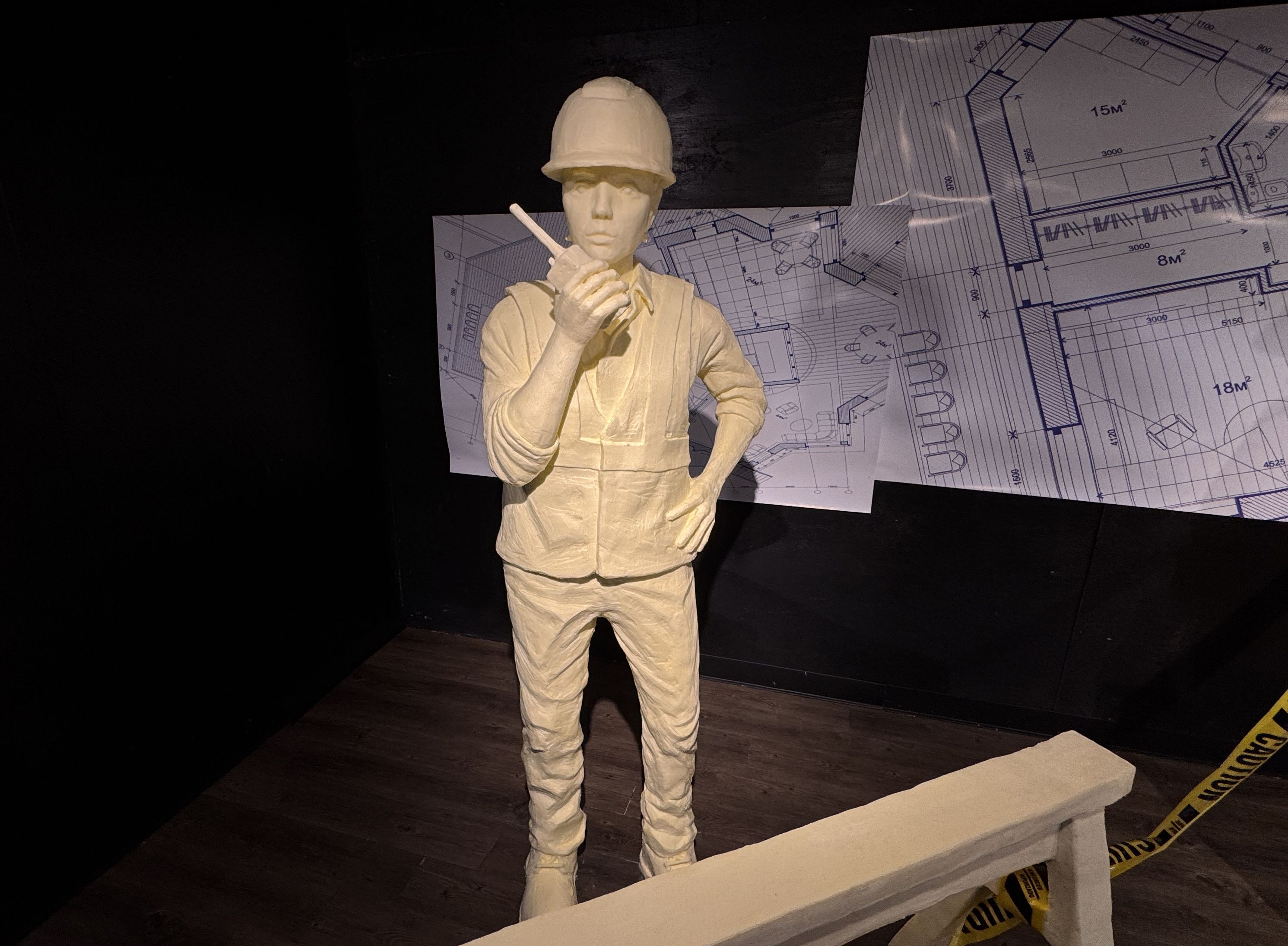 Butter sculpture of a construction worker in a hard hat and vest holding a walkie-talkie, standing in front of architectural blueprints on a black wall with caution tape on the floor.