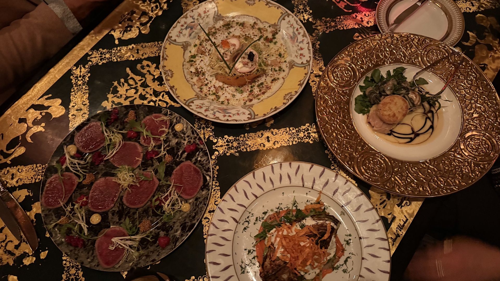 Overhead view of an ornate dinner table with four plates: a black marble tuna carpaccio with sprouts; a dish with ringed sauce and chives; a gold charger with scallops and greens; a white plate with vegetables.