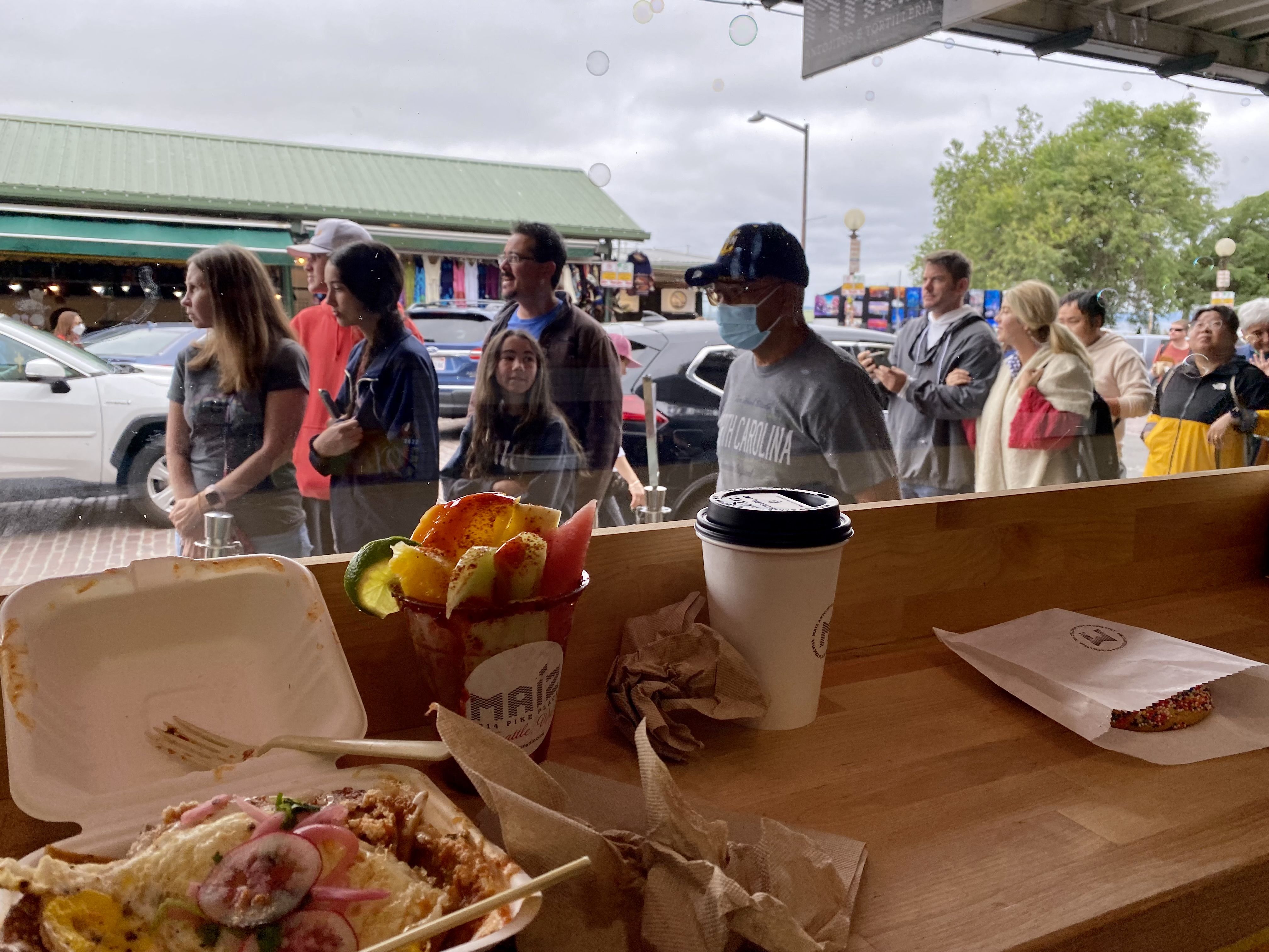 A counter with messy napkins, a to-go container of chilaquiles and a fruit cup, with a window showing a view of people waiting outside.
