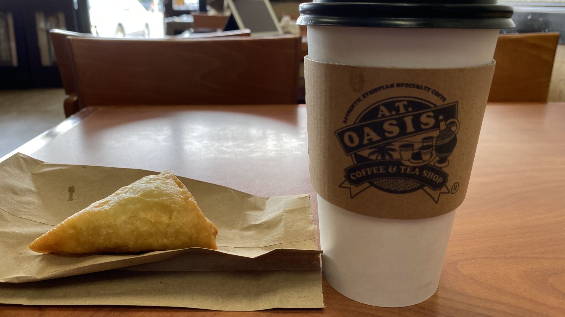 A coffee cup with a cardboard sleeve reading AT Oasis Coffee and tea on a table next to a triangular pastry