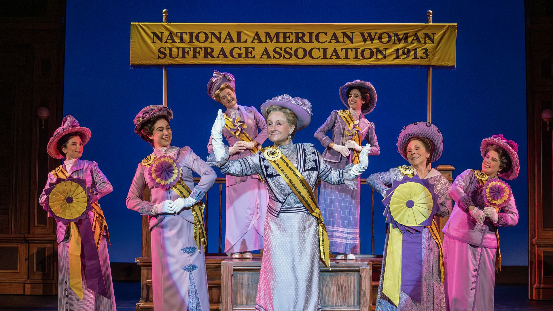 Seven women in early 1900s-style dresses and hats stand under a yellow banner reading "National American Woman Suffrage Association 1913." They wear yellow sashes and hold ribbons.