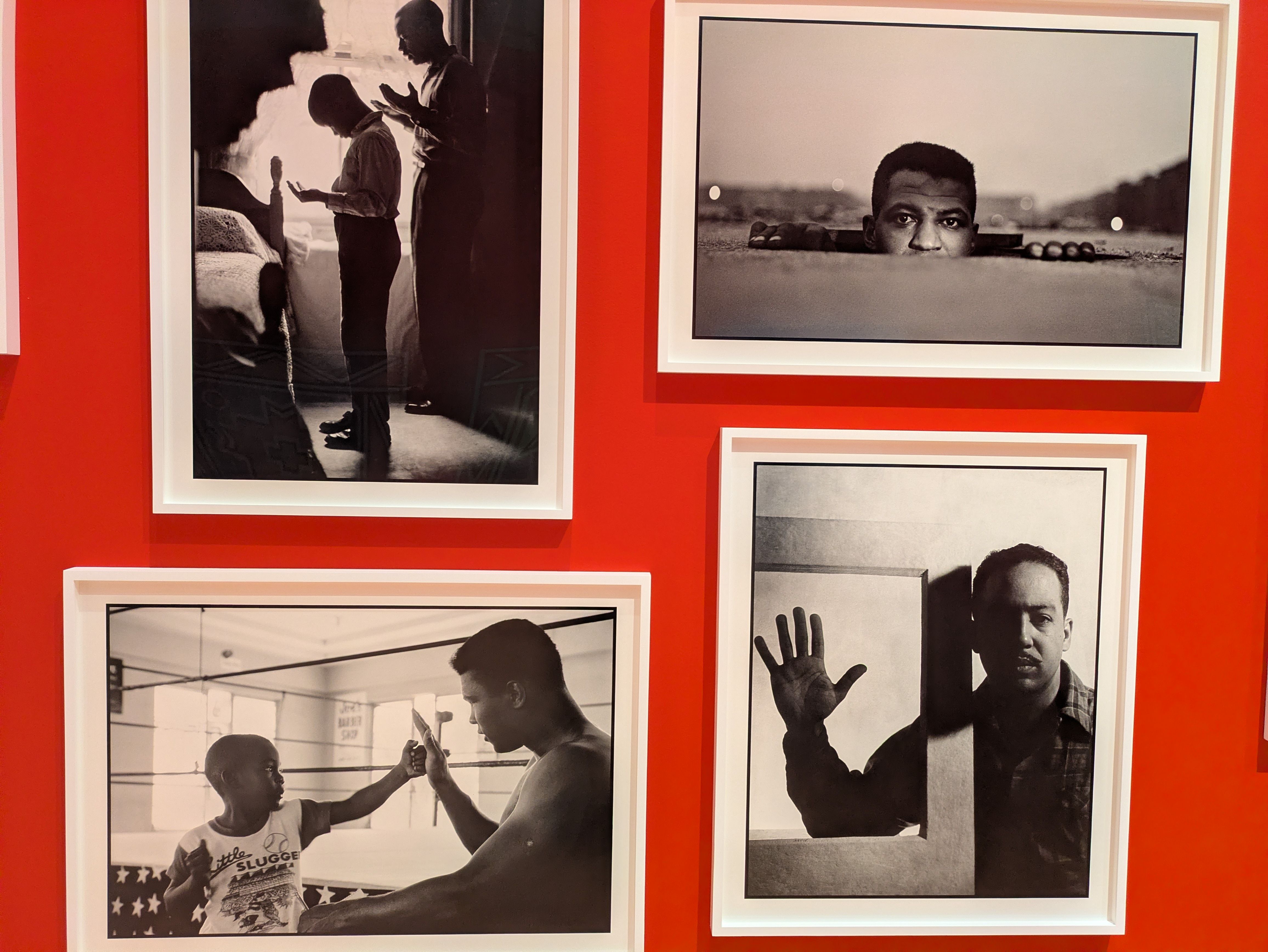 Black-and-white photographs framed on a vivid red gallery wall; left image shows silhouettes of people in a room, right image shows a man peeking over a surface.