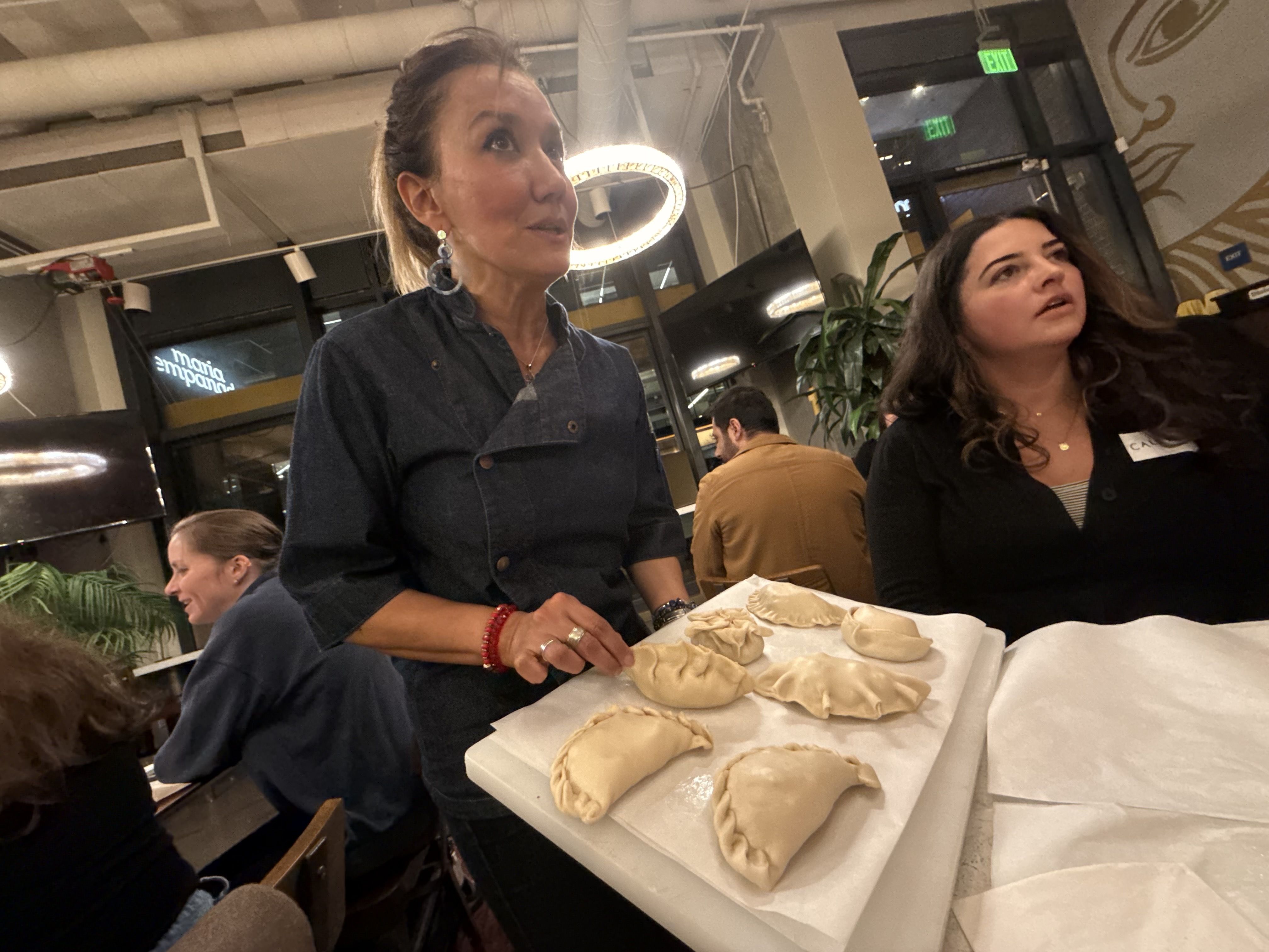 A woman in a dark chef's coat shows a tray of uncooked empanadas in a bustling restaurant with diners in the background and circular ceiling lights.