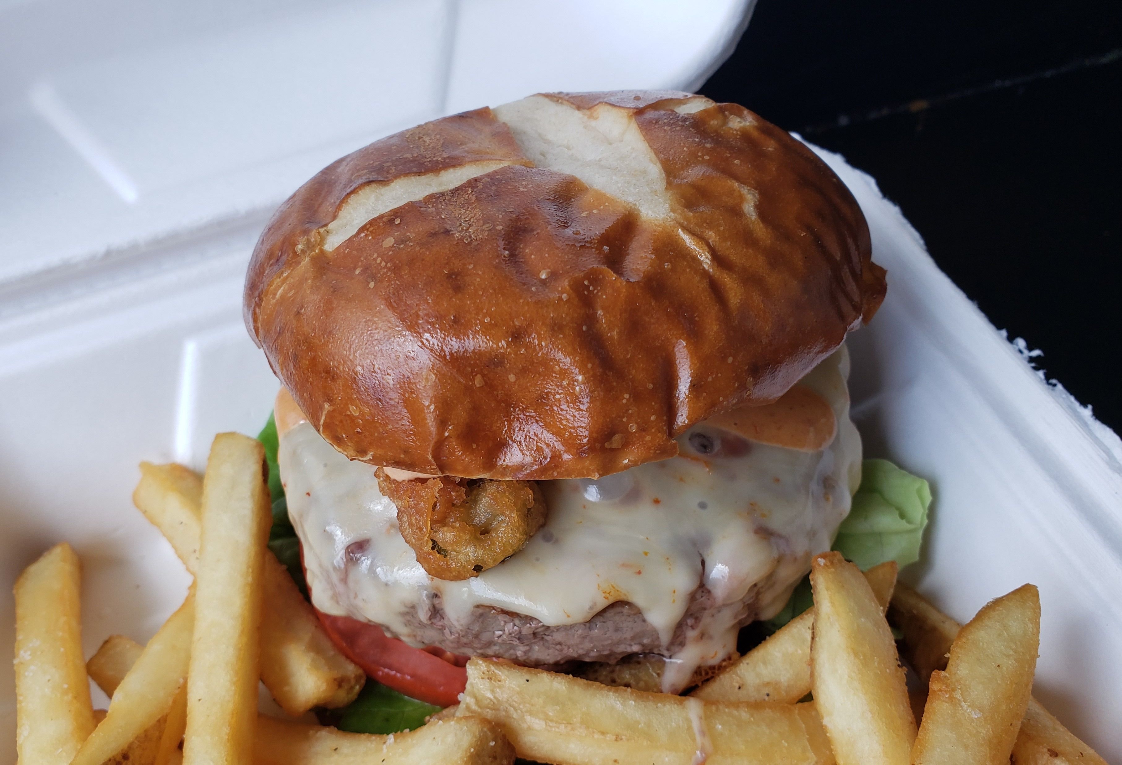 Burger with melted cheese on pretzel bun, surrounded by French fries in a styrofoam box.