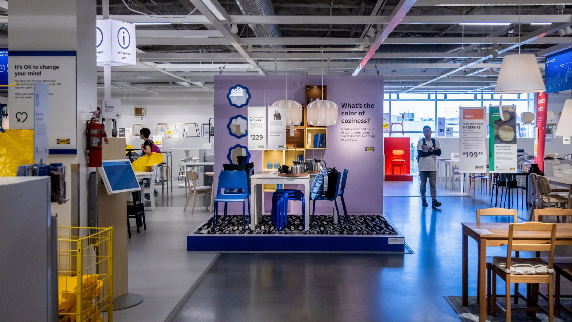 Interior of an IKEA store showing displayed furniture sets with price tags, people walking, and bright lighting under an industrial ceiling.
