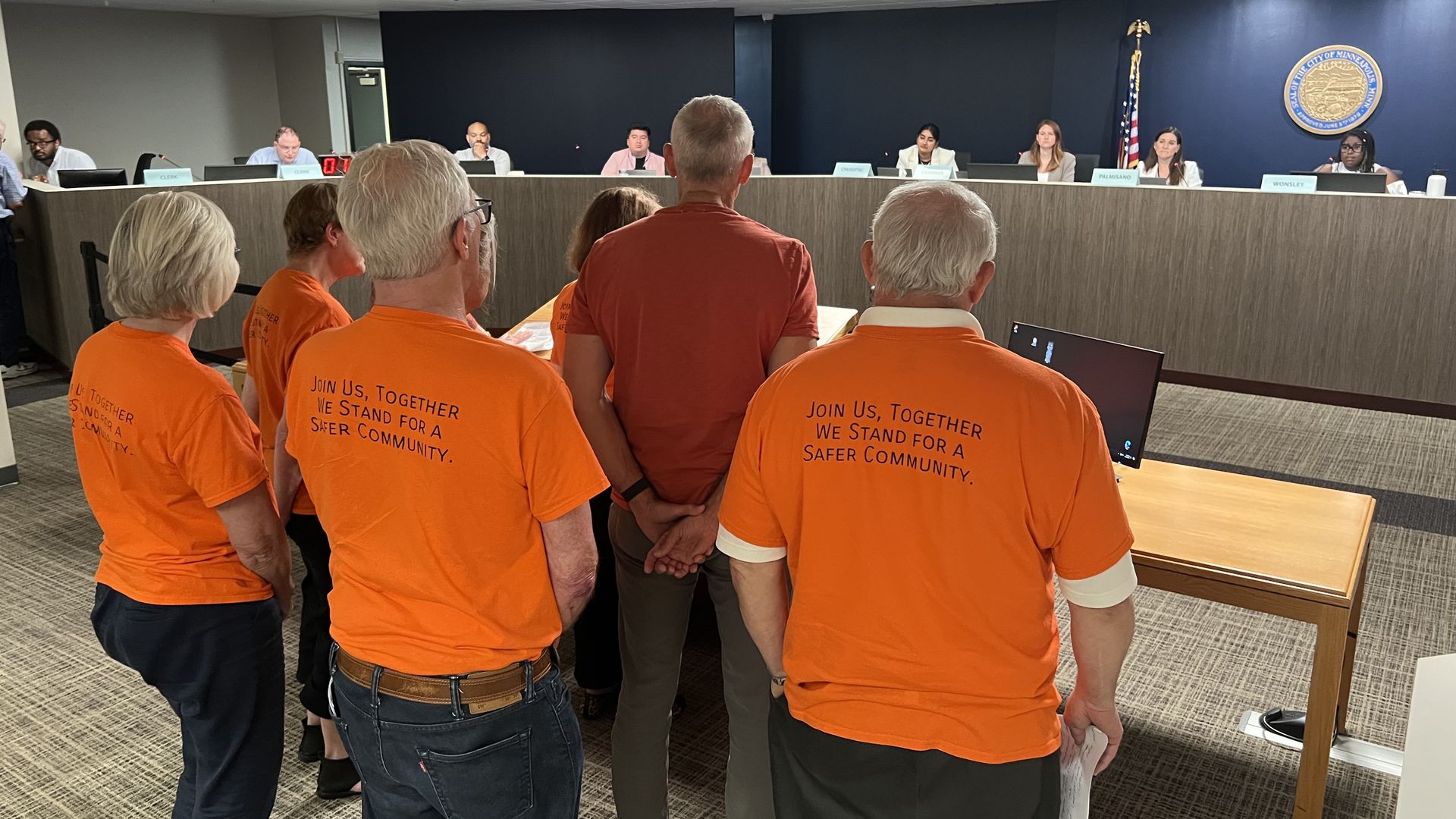 Several people stand facing away from the camera and toward a dais with Minneapolis City Council members. The public commenters wear orange shirts.