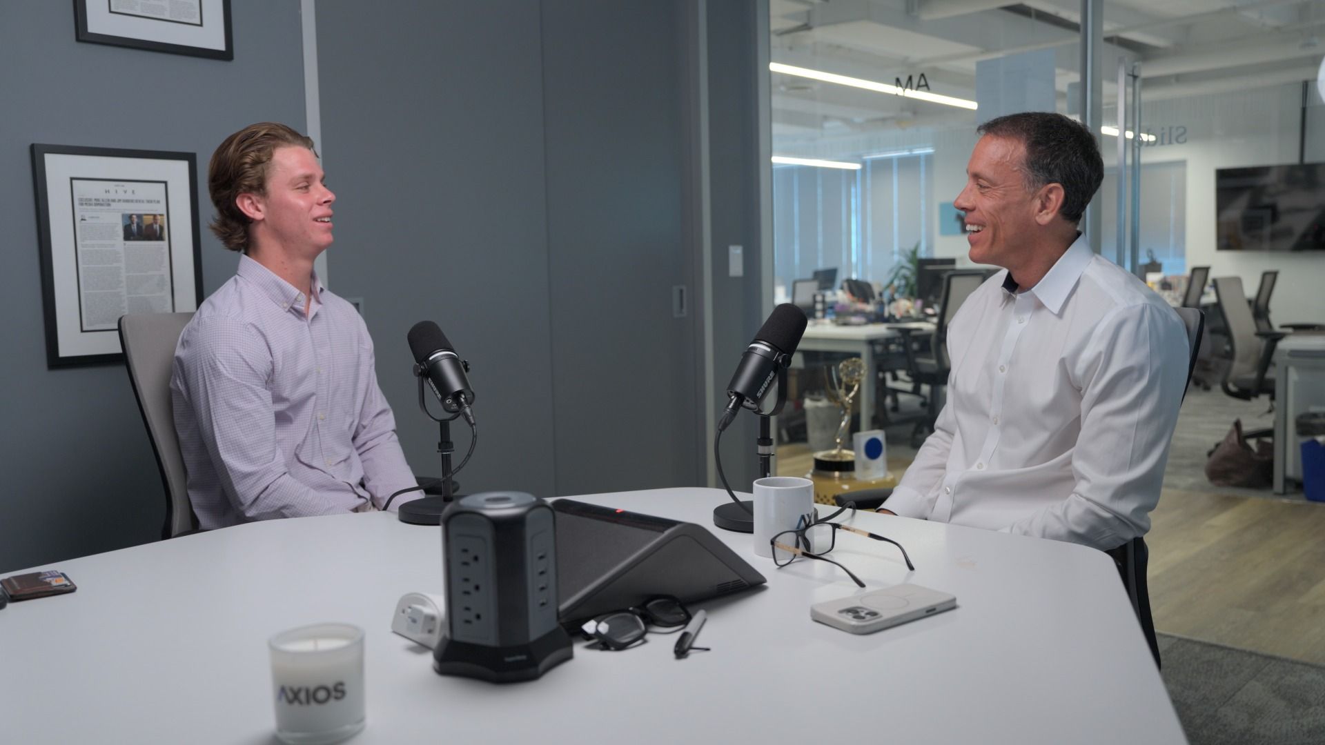 Two men in a glass-walled office podcast studio sit across a white table with black microphones, talking; desks and chairs visible in the background, and a mug labeled AXIOS on the table.