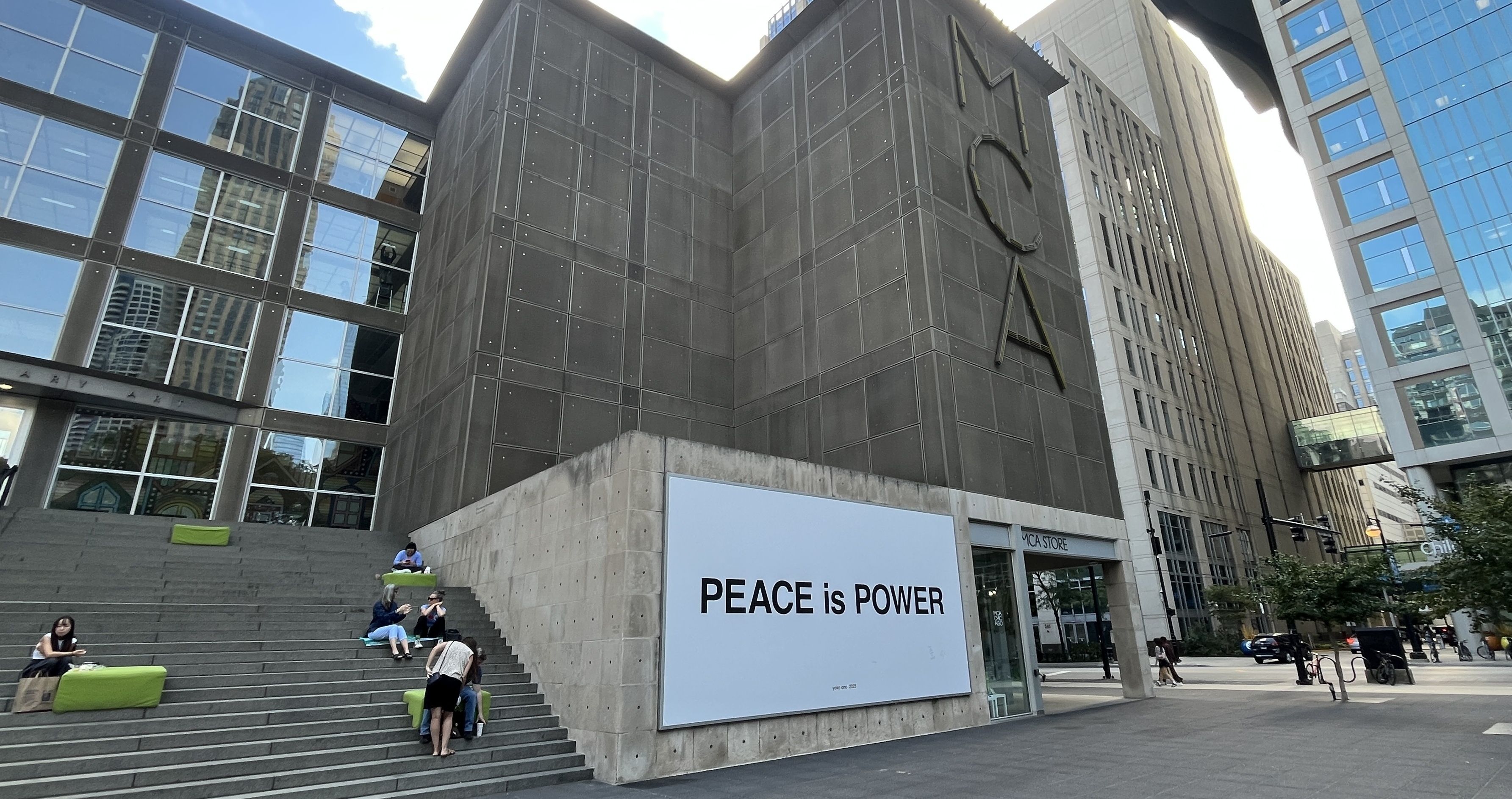 Outdoor stairs by a modern building with large glass windows, several people sitting on green cushions, and a large wall sign saying "PEACE is POWER" in black letters on white background.