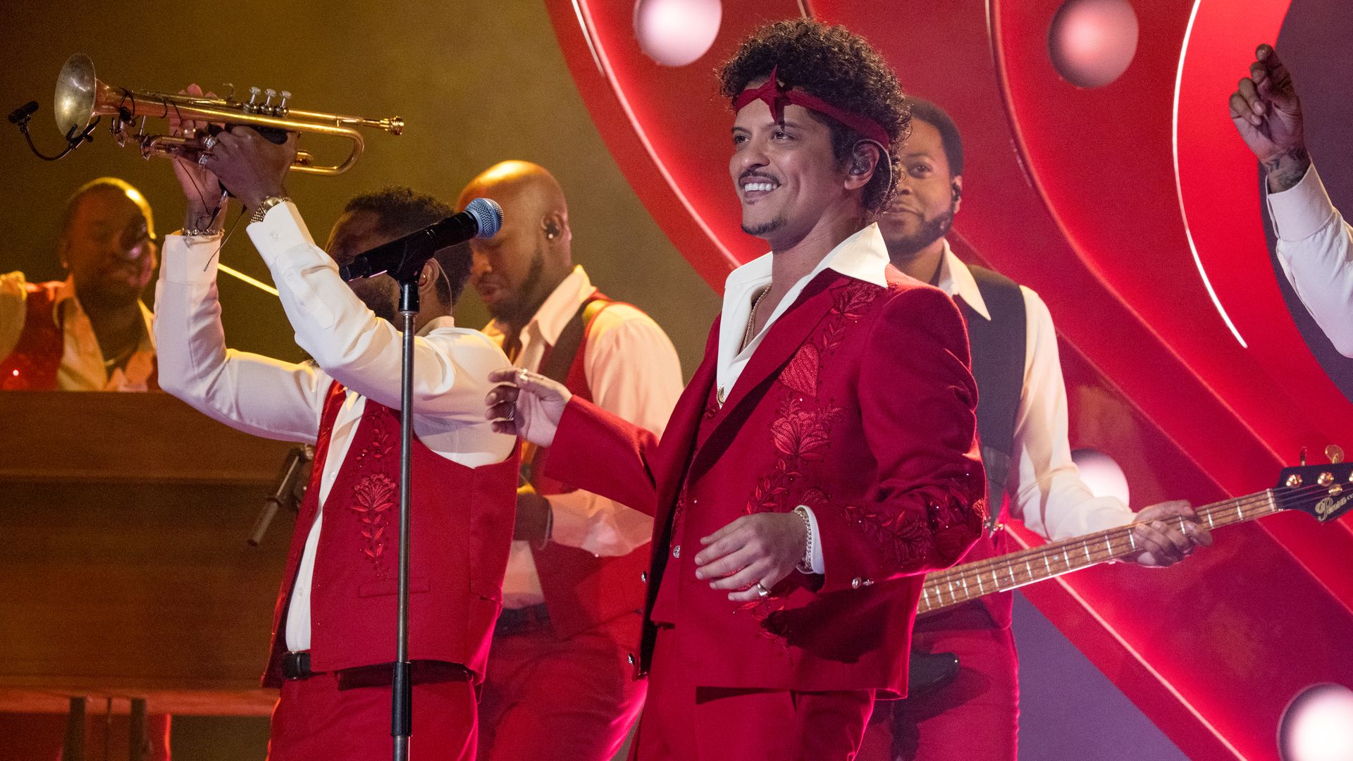 Live stage performance: a man in a red embroidered suit sings into a microphone, wearing a red headband; red-clad band members, trumpet raised, with a bold red circular backdrop.