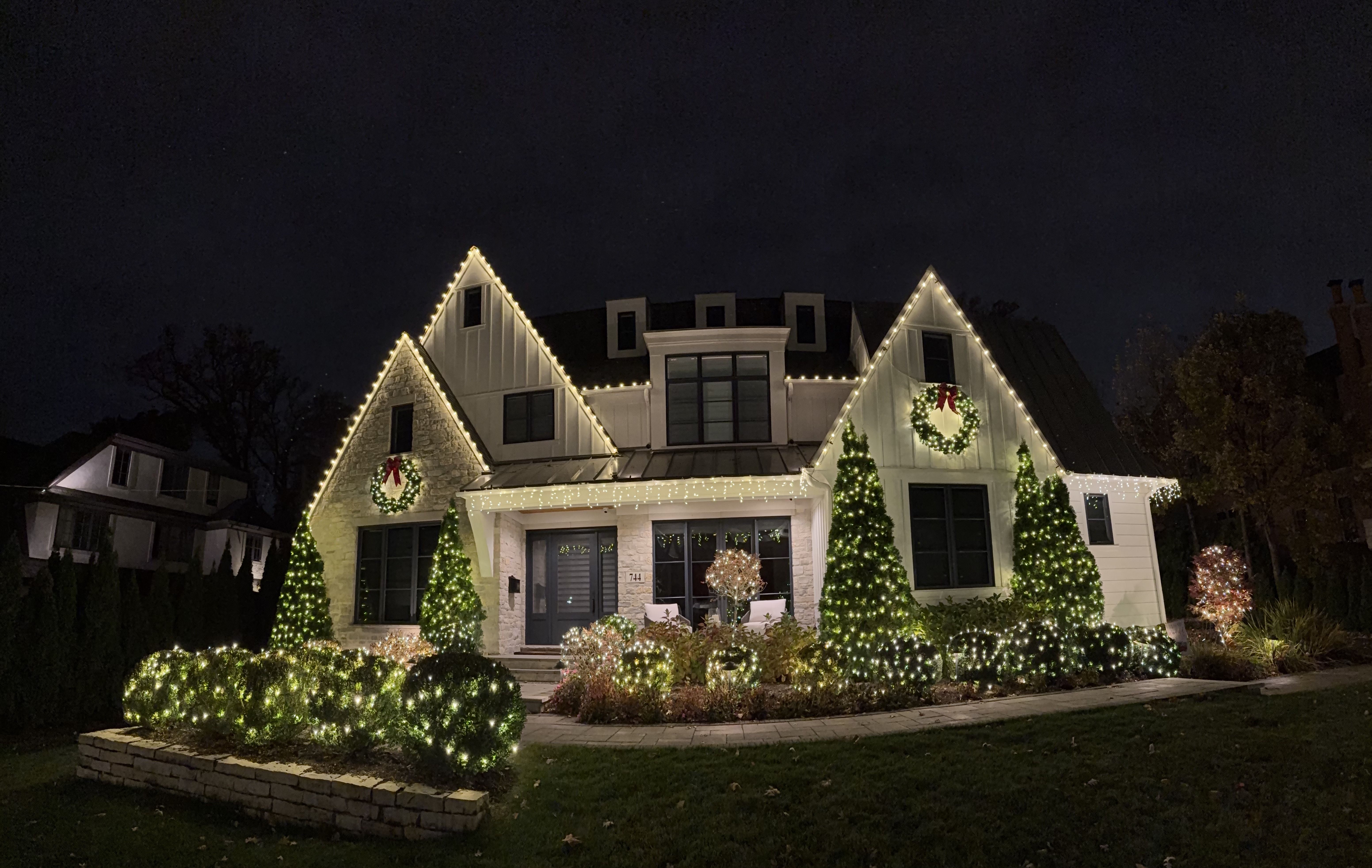 A large house decorated with white Christmas lights outlining the roof, windows, and bushes. Two wreaths with red bows hang on the front, and trees are wrapped in lights.