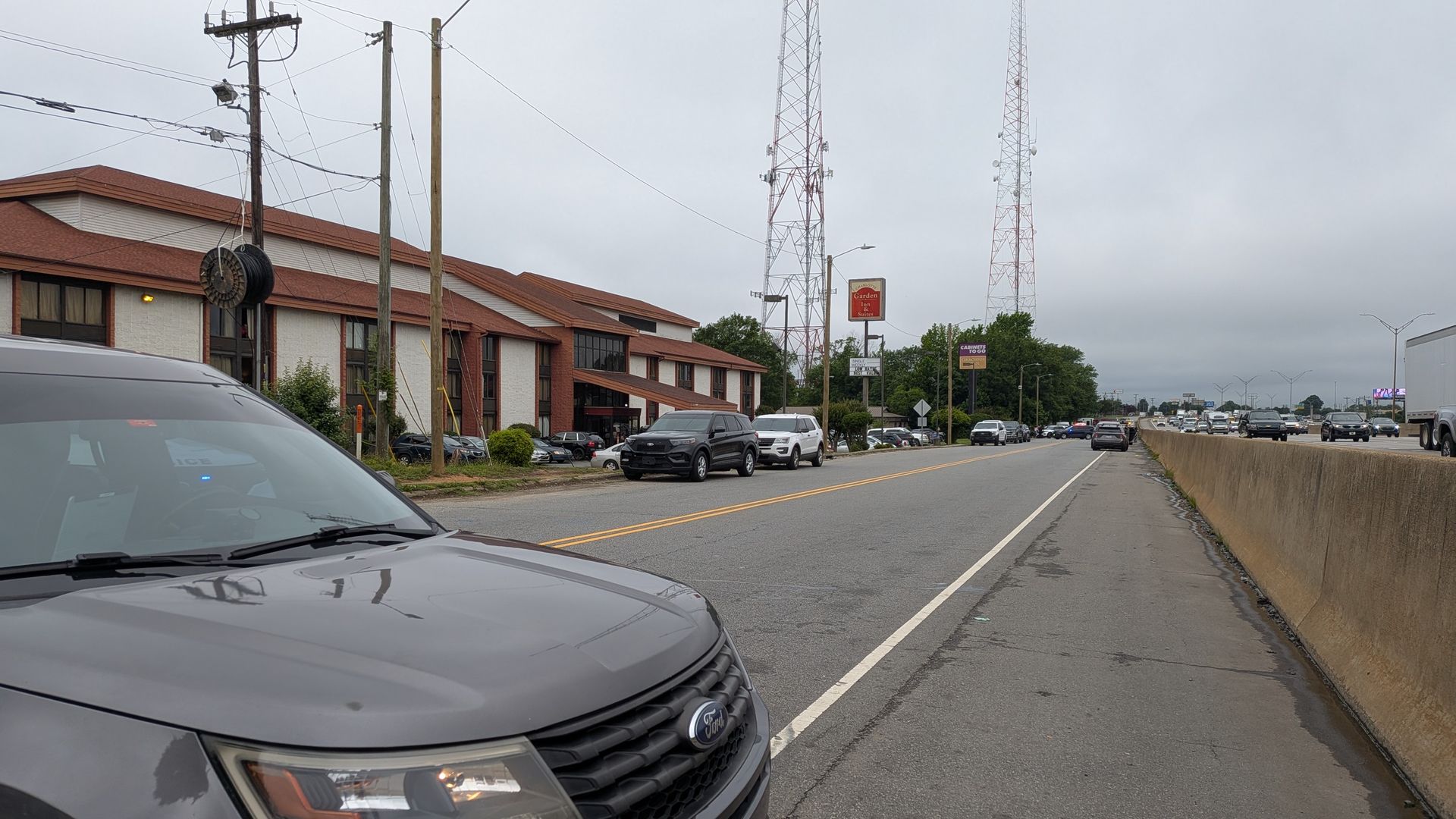 Overcast street scene with the hood of a gray Ford in the foreground. Left: red-tile motel, utility poles and a large cable reel. Center: two tall radio towers and a Gardens Inn Suites sign; traffic on the road.
