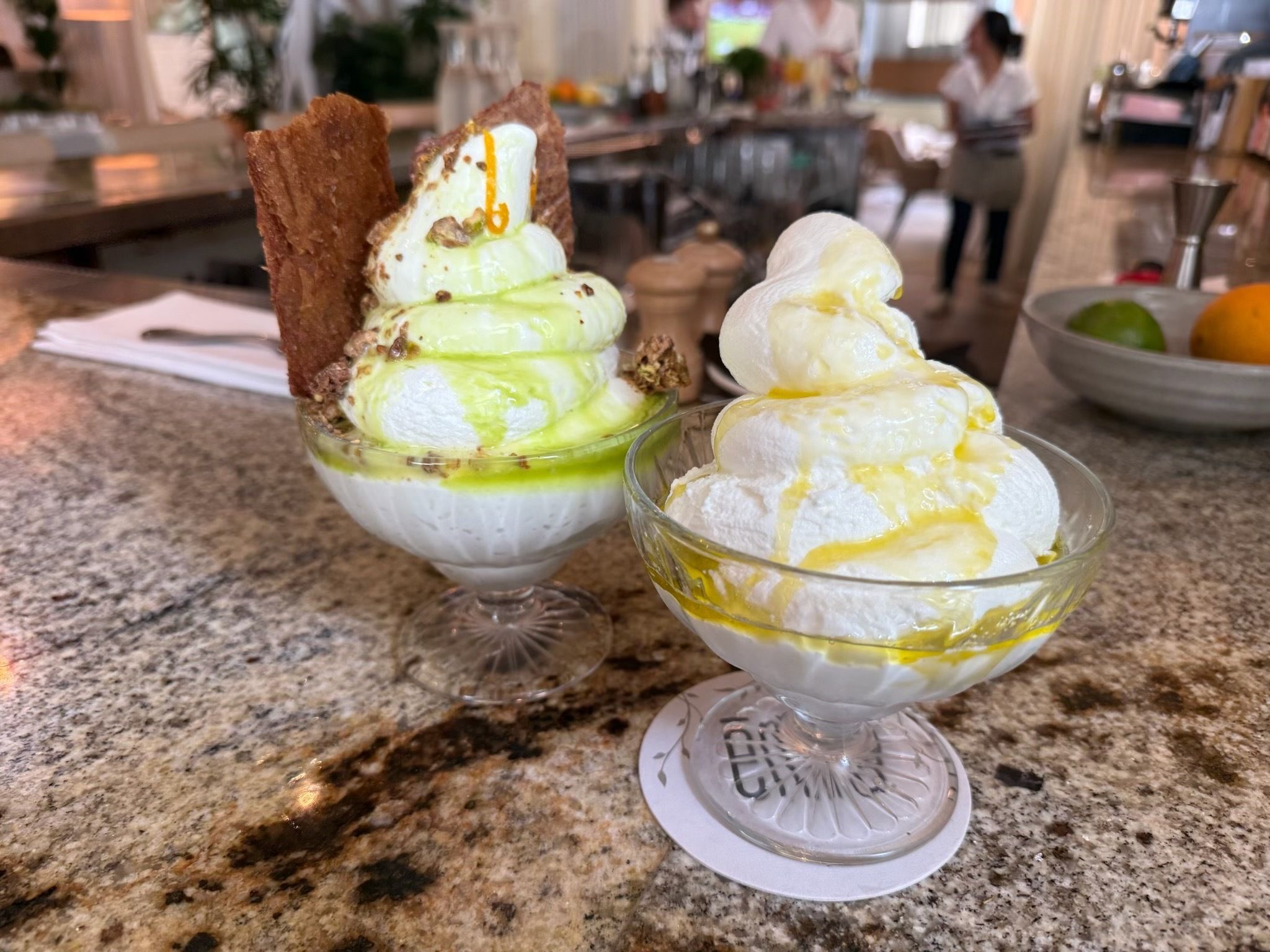 Two ice cream sundaes in glass bowls on a speckled counter: pistachio-green and vanilla scoops with a chocolate shard, pistachio drizzle and lemon glaze, blurred cafe scene in the background.