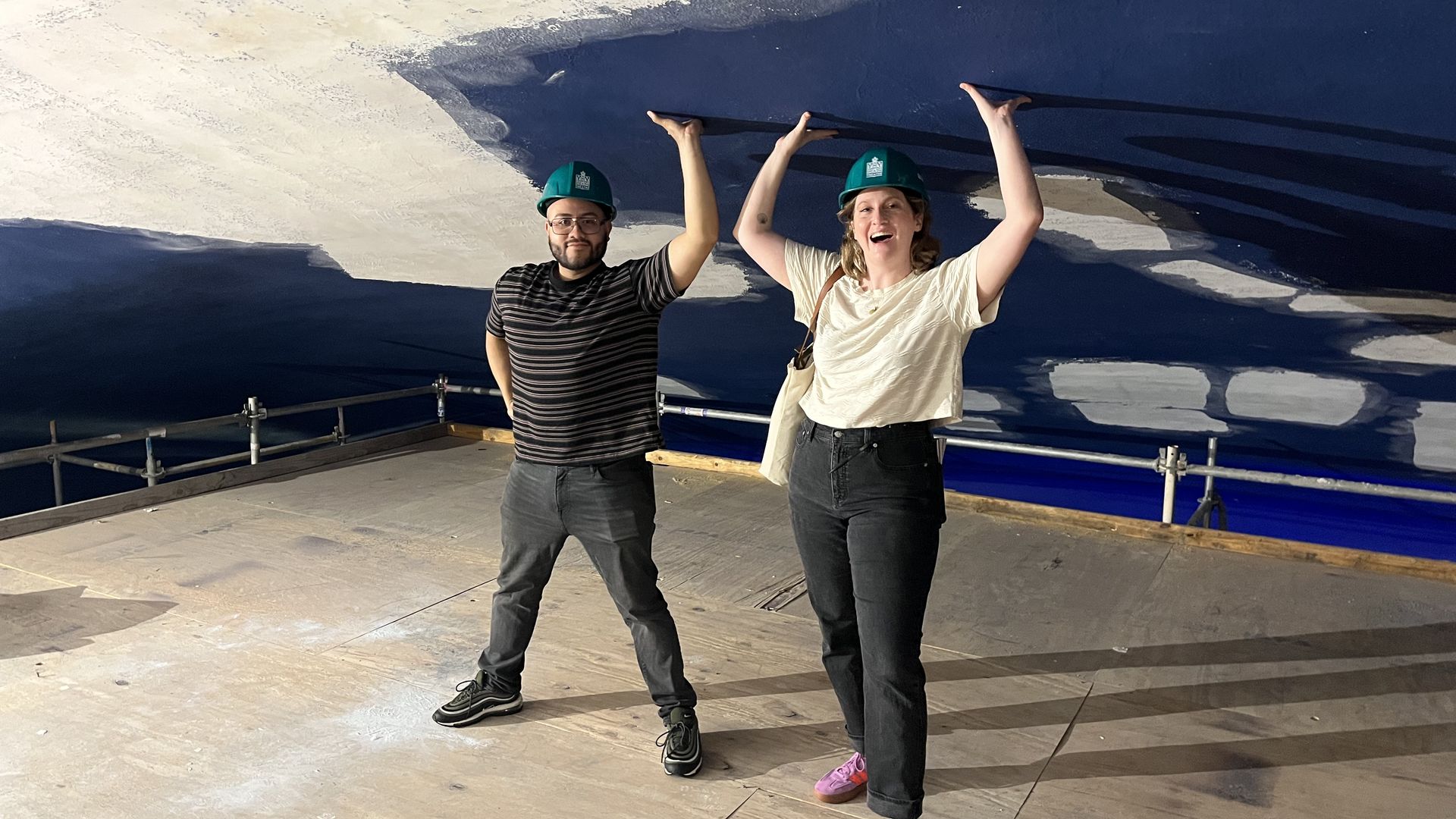 Two people wearing green hard hats smile and raise their arms inside a construction area, under a blue ceiling with white patches; metal railing and a wooden floor are visible.